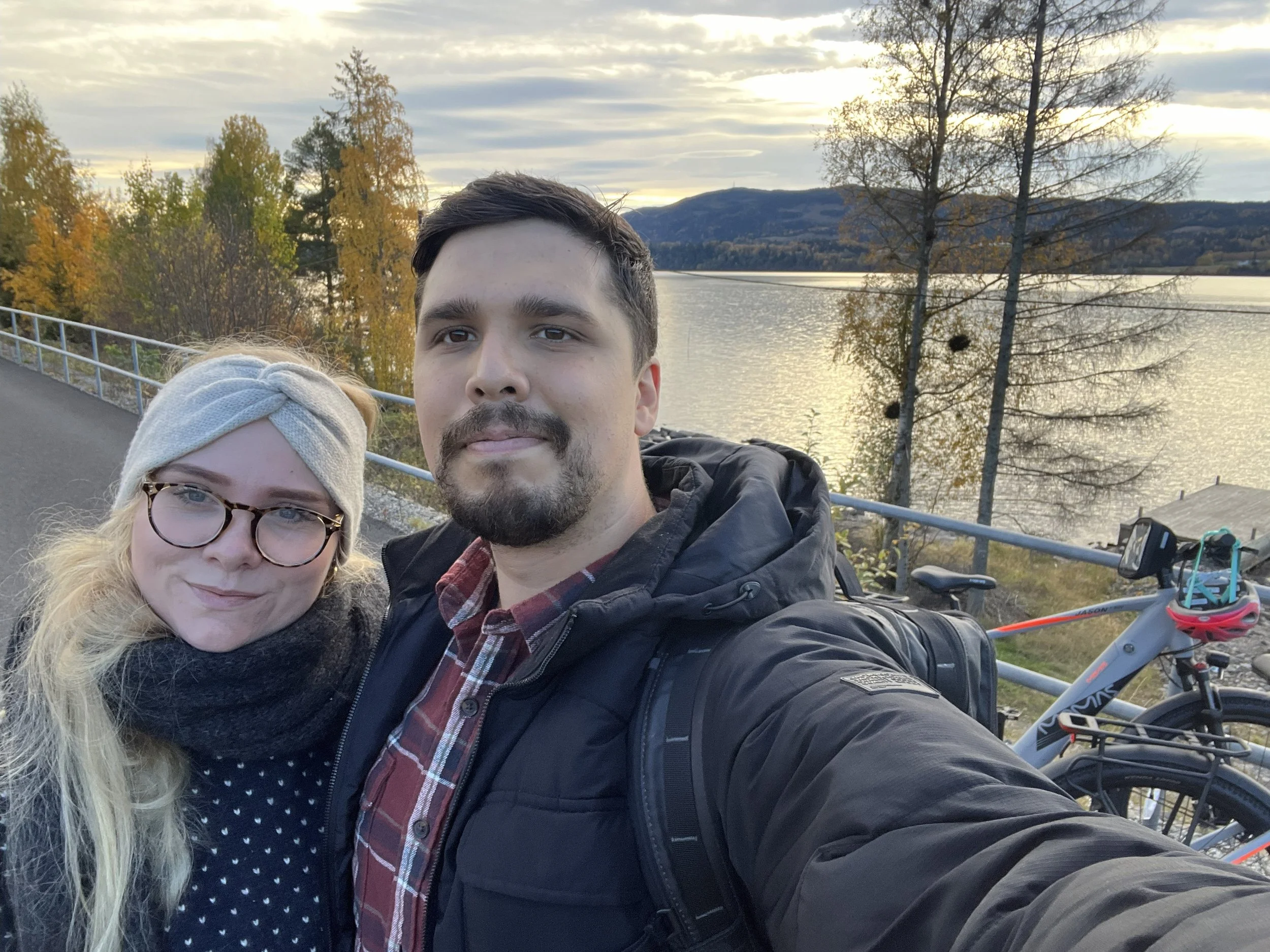 A couple taking a selfie outdoors near a body of water surrounded by trees with autumn foliage, mountains in the background, and two bicycles nearby.