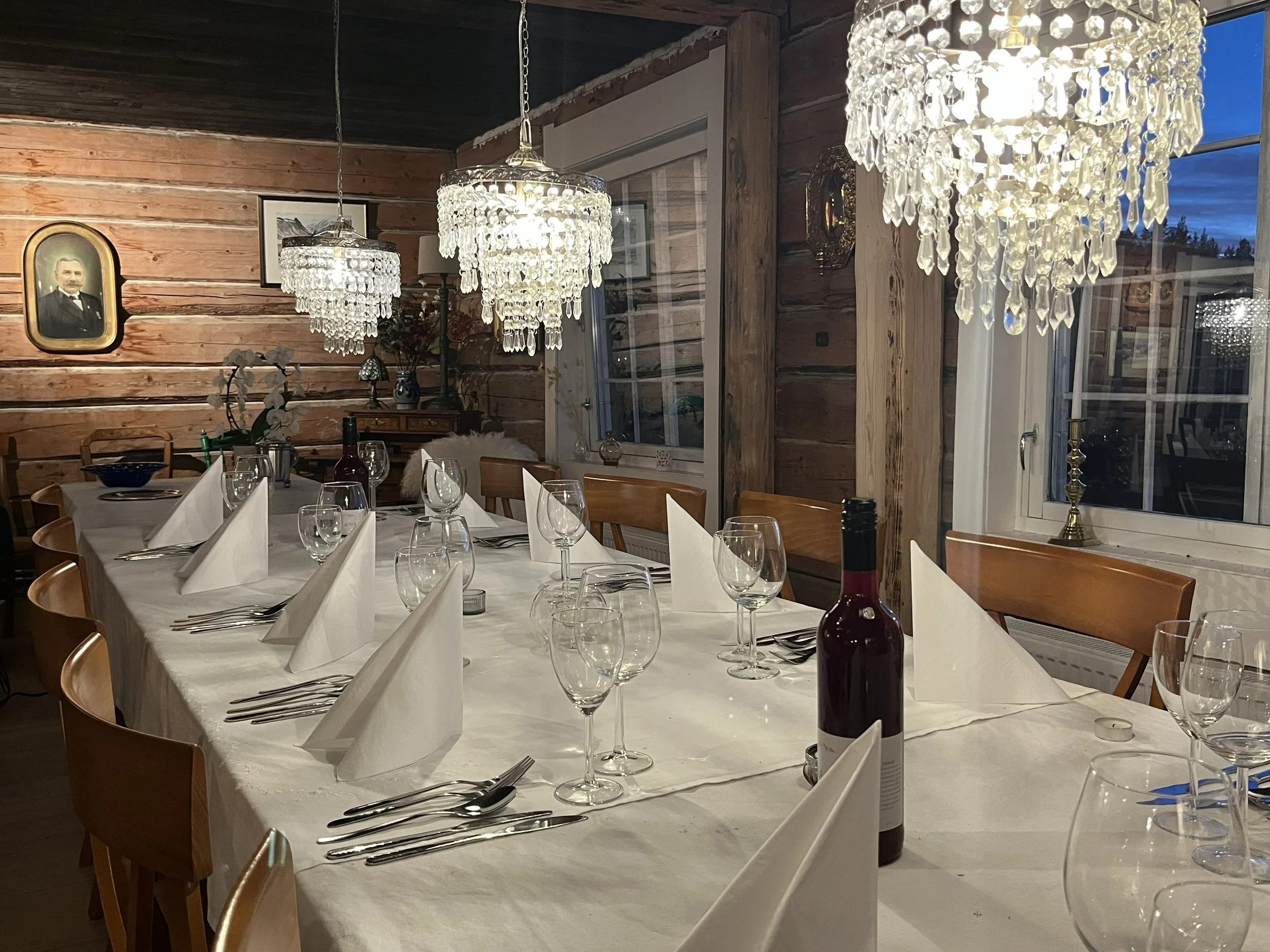 A dining table set for a meal with white napkins, wine glasses, and cutlery, illuminated by chandeliers in a rustic wood-paneled room with windows showing a dusk sky.