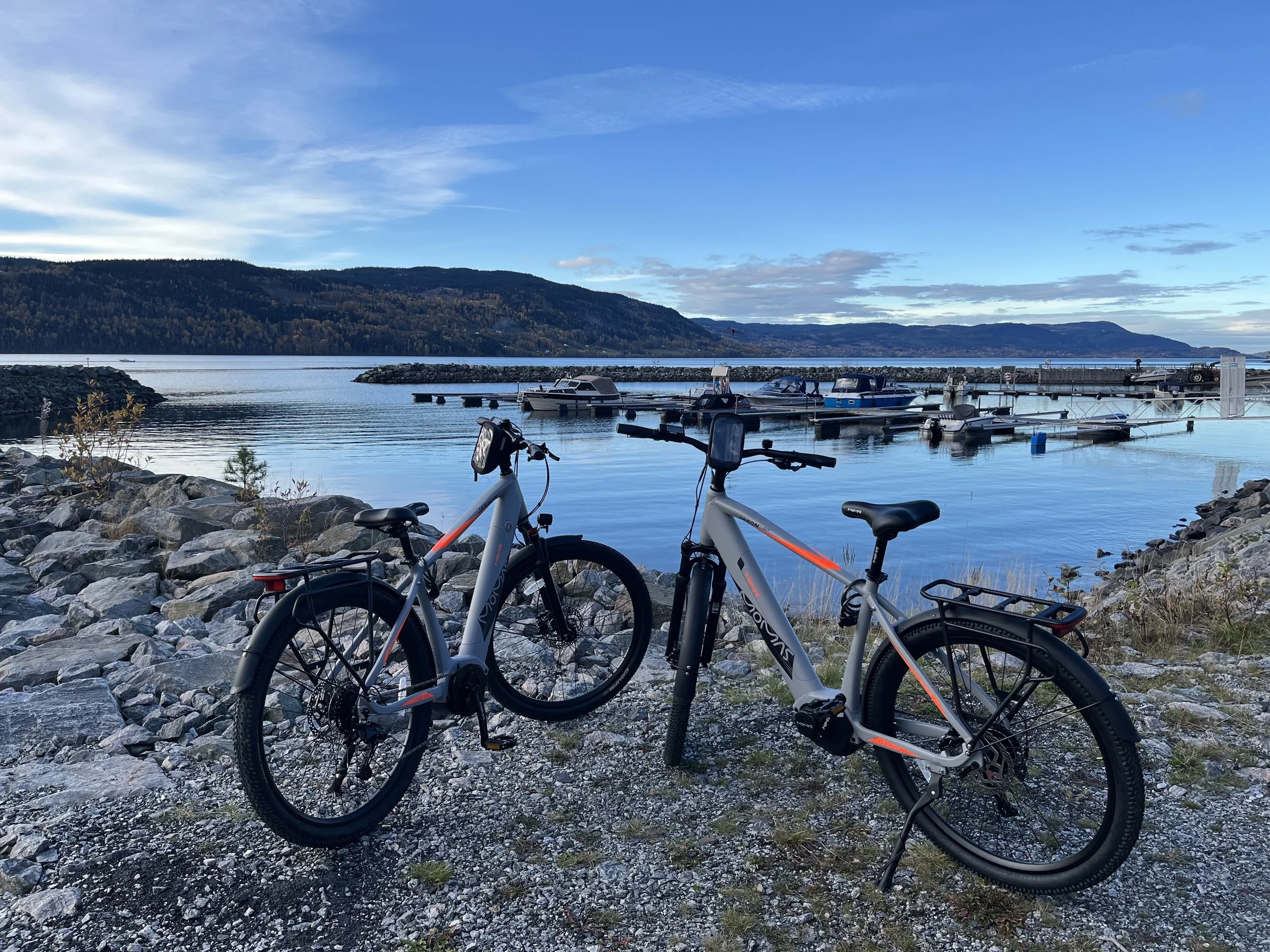 Two electric bikes on a rocky shore next to a lake with boats docked and hills in the background under a blue sky with some clouds.