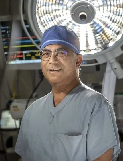 Dr. Uttam Sinha in scrubs and surgical cap standing in an operating room with medical equipment and surgical light overhead.