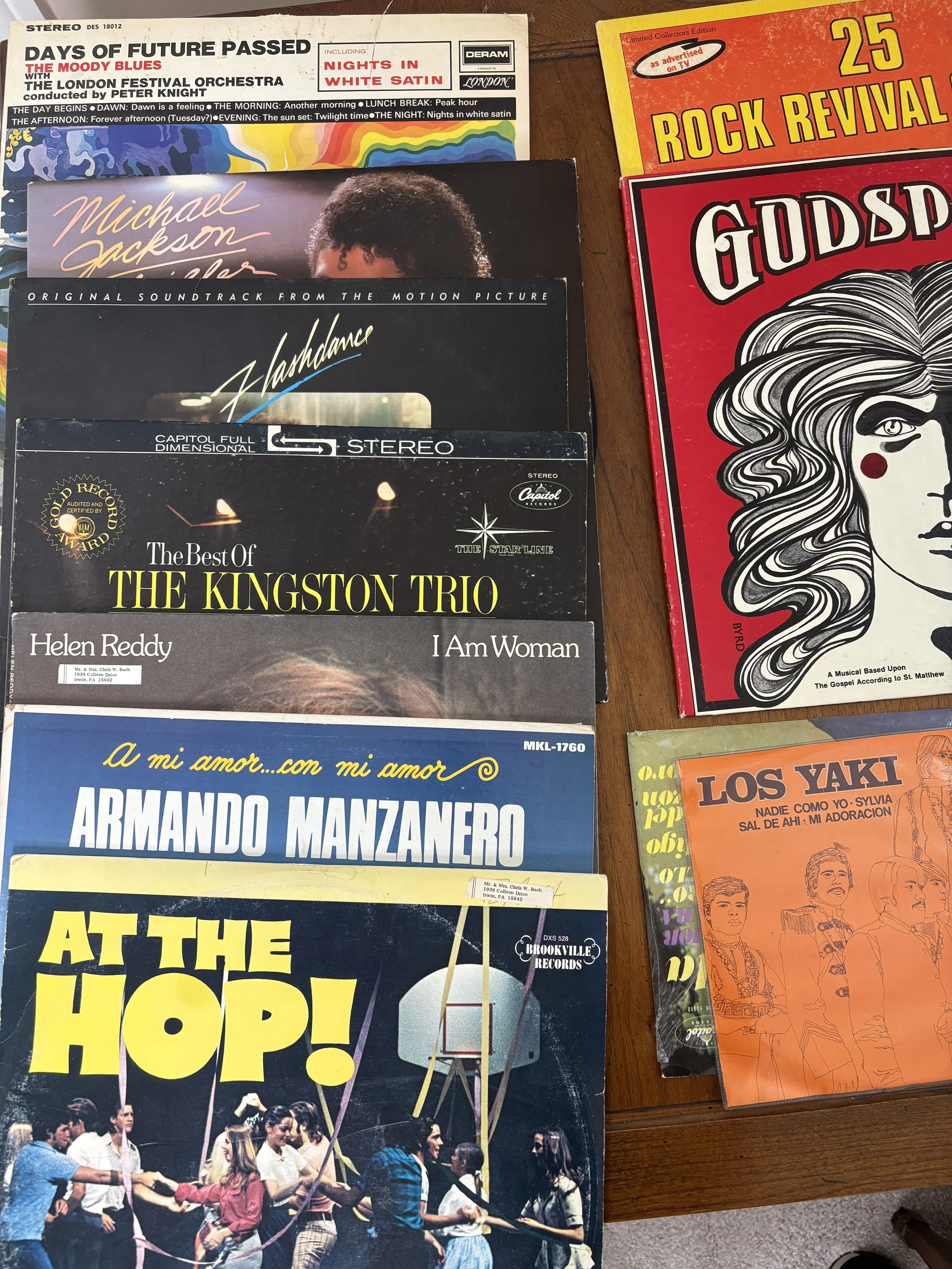 Collection of vintage vinyl records and album covers on a wooden table, including 'Days of Future Passed' by The Moody Blues, 'Michael Jackson Thriller,' 'Handclap,' 'The Best of The Kingston Trio,' 'I Am Woman' by Helen Reddy, 'A mi amor... con mi a