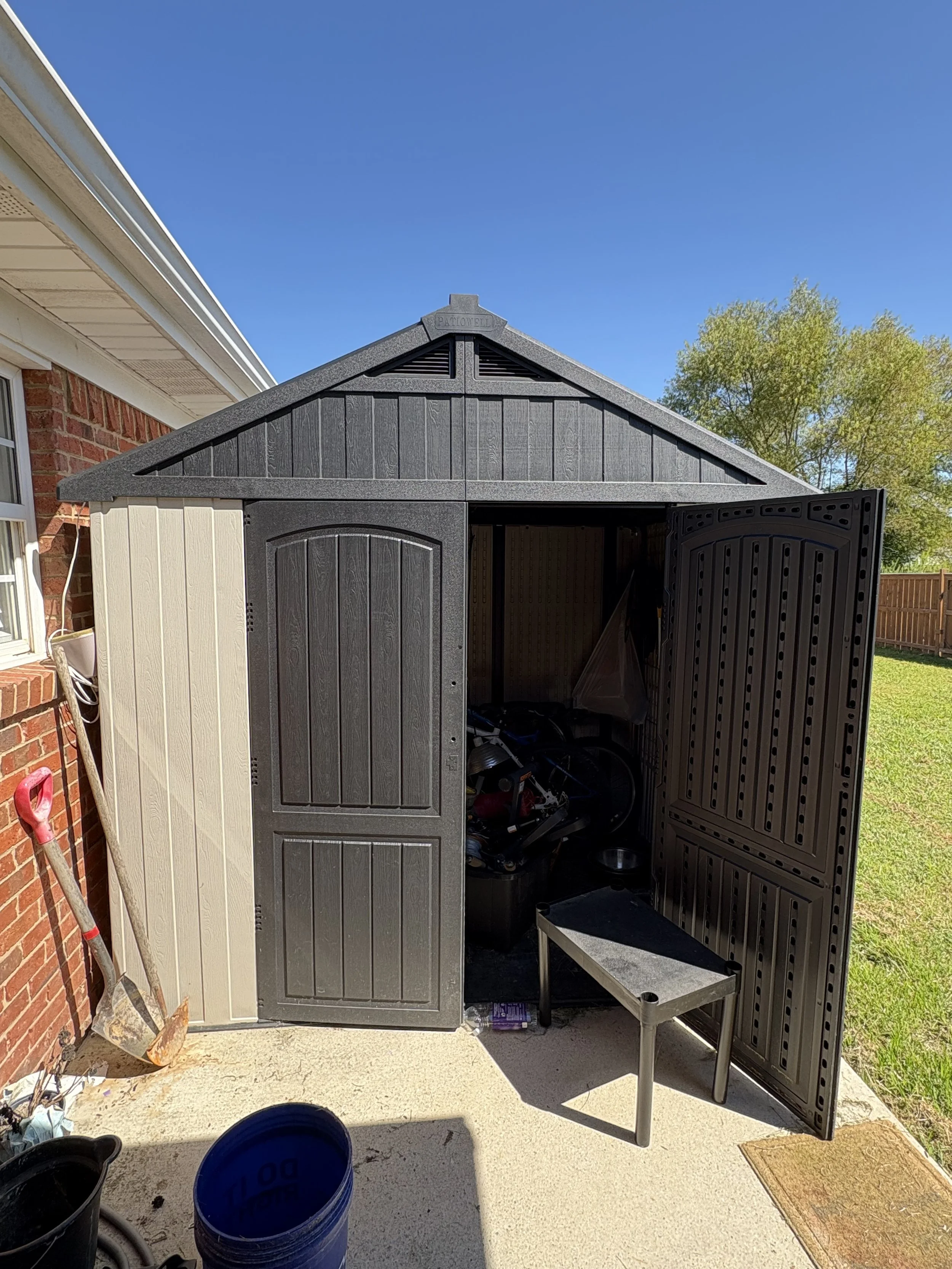 An outdoor shed with black and beige doors, open to reveal tools and equipment inside, located next to a brick house on a concrete patio with a shadow cast on the ground.