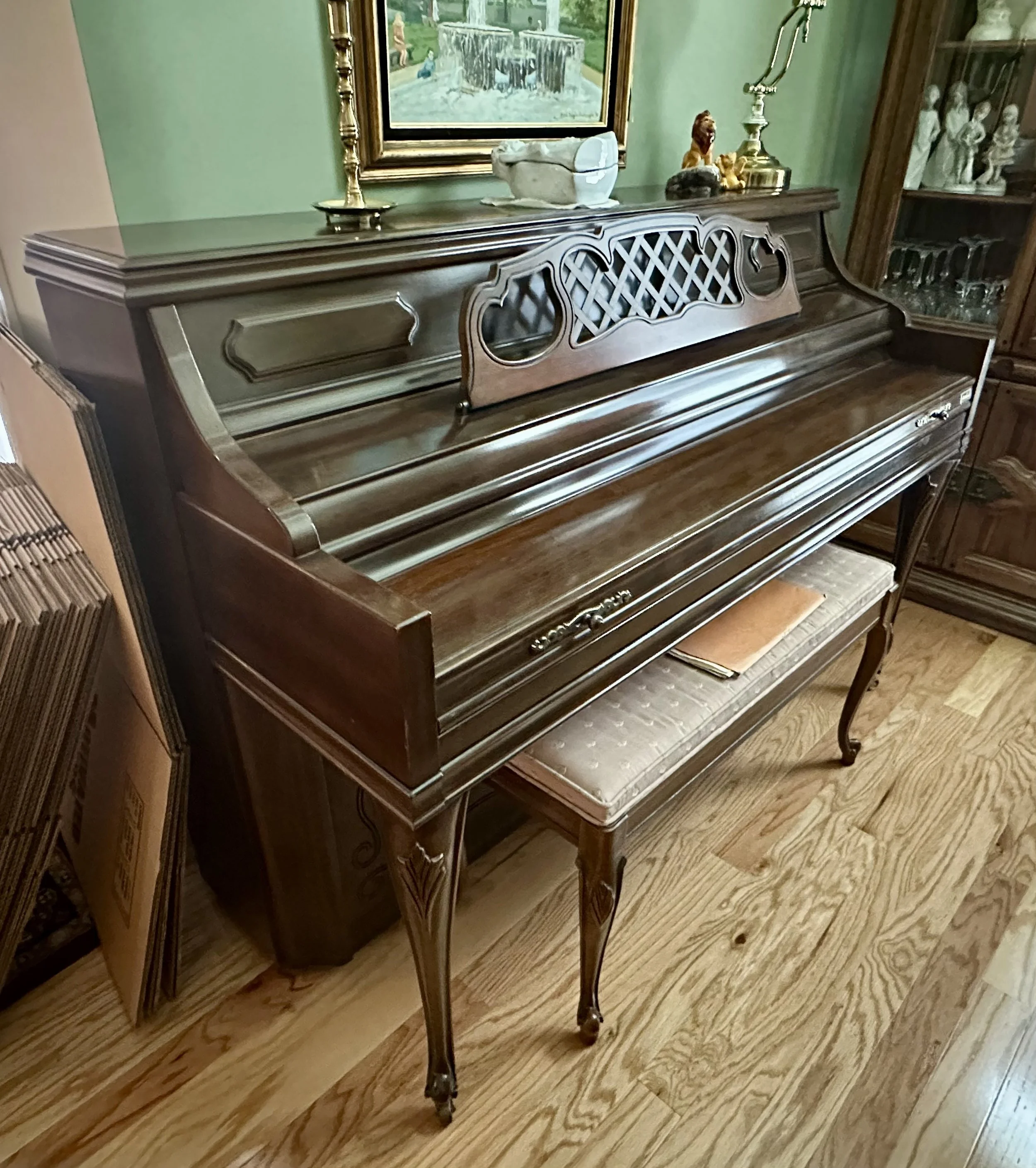 An antique upright piano with a wooden finish positioned against a green wall, with a cushioned bench underneath. The piano has a decorative music stand and is located in a room with wooden flooring.