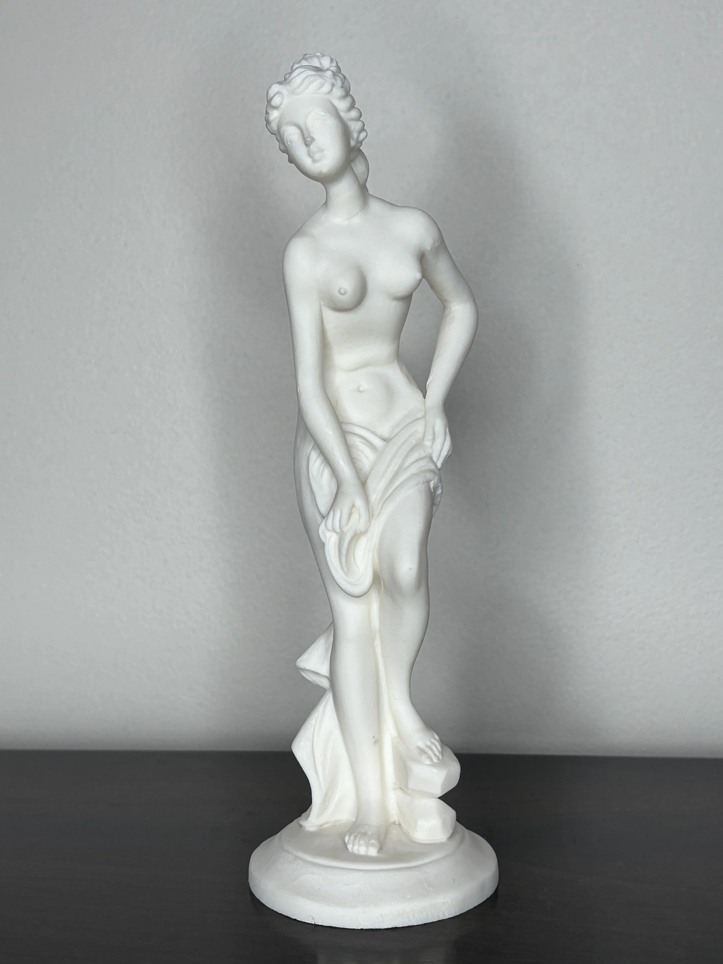 White sculpture of a woman standing with her hair styled in curls, holding a piece of cloth near her waist, on a round base.