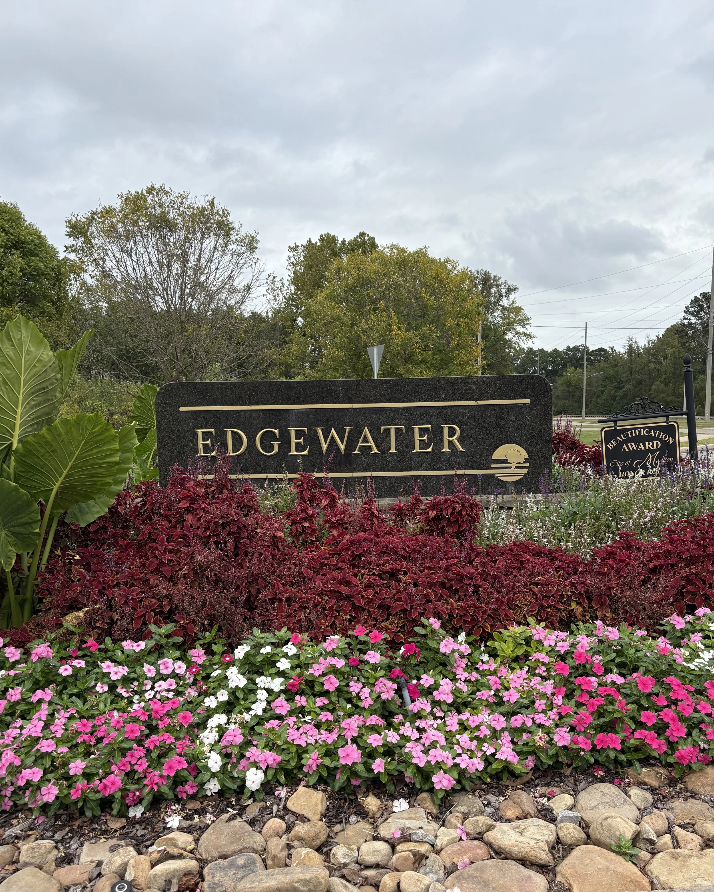 Entrance sign for Edgewater with colorful flowers and greenery in the foreground, overcast sky in the background.
