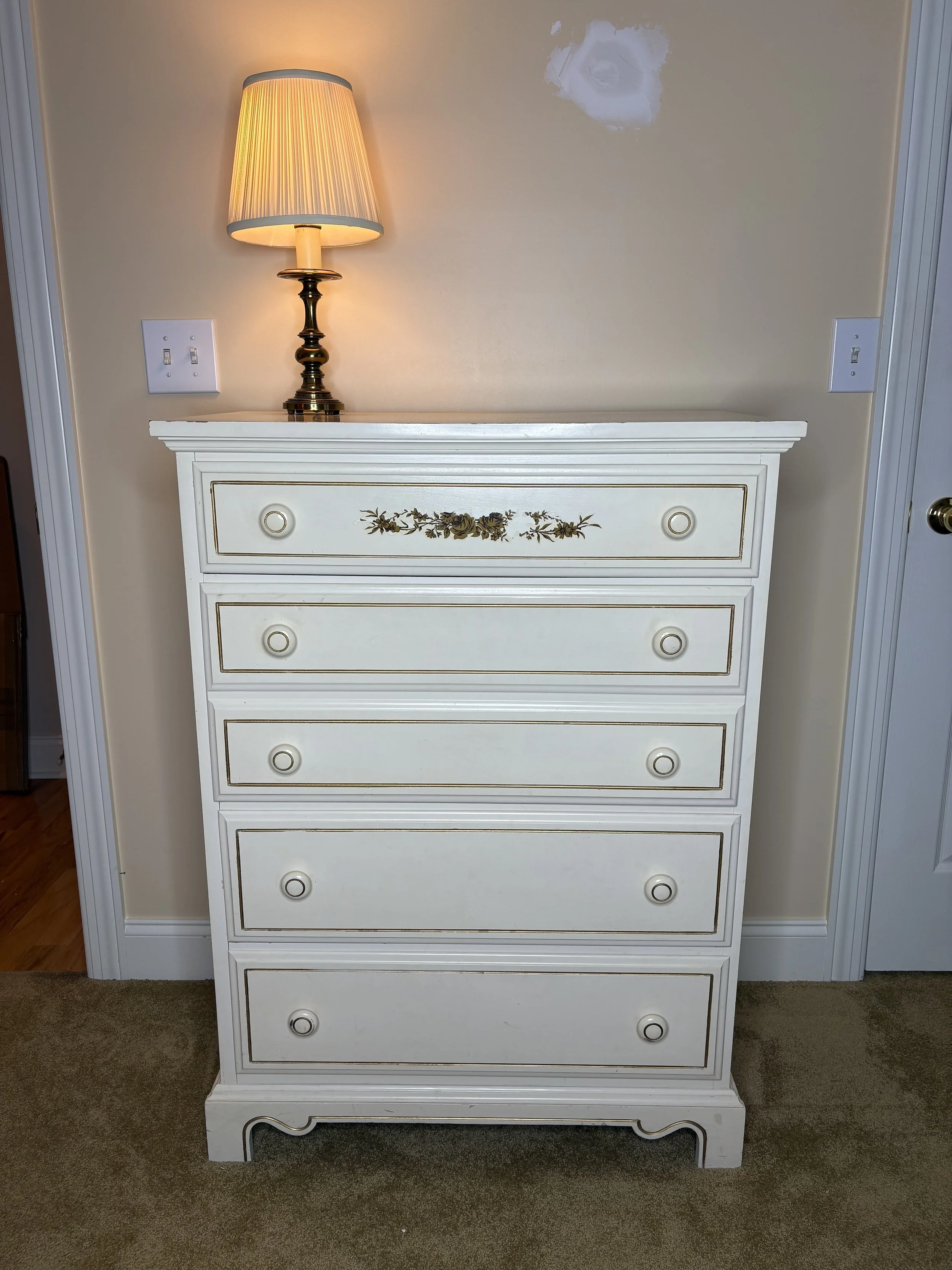 White dresser with five drawers and gold accents, topped with a table lamp with a beige shade, on a beige wall with a hole or damage in the wall behind the lamp.