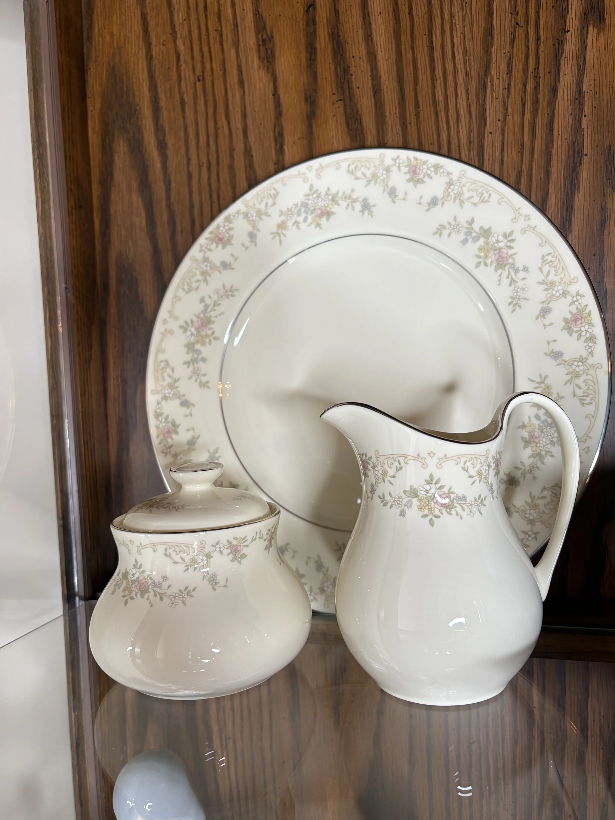 A porcelain tea set consisting of a teapot with a lid, a creamer, and a large dinner plate, all with a floral pattern featuring pink and green flowers on a white background, displayed on a wooden surface.