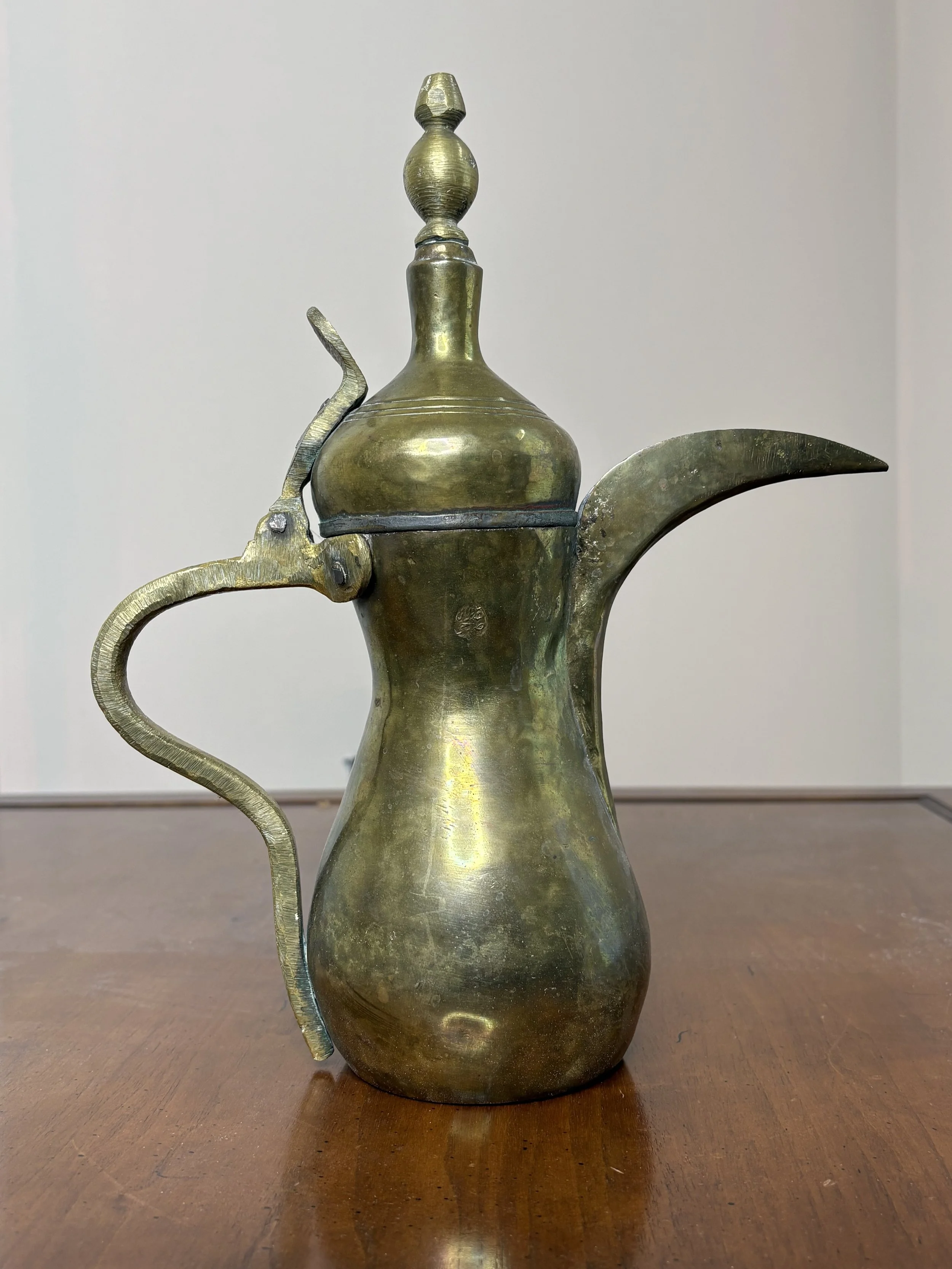 An antique brass teapot with a long, curved spout, a handle, and an ornate lid, sitting on a wooden surface.