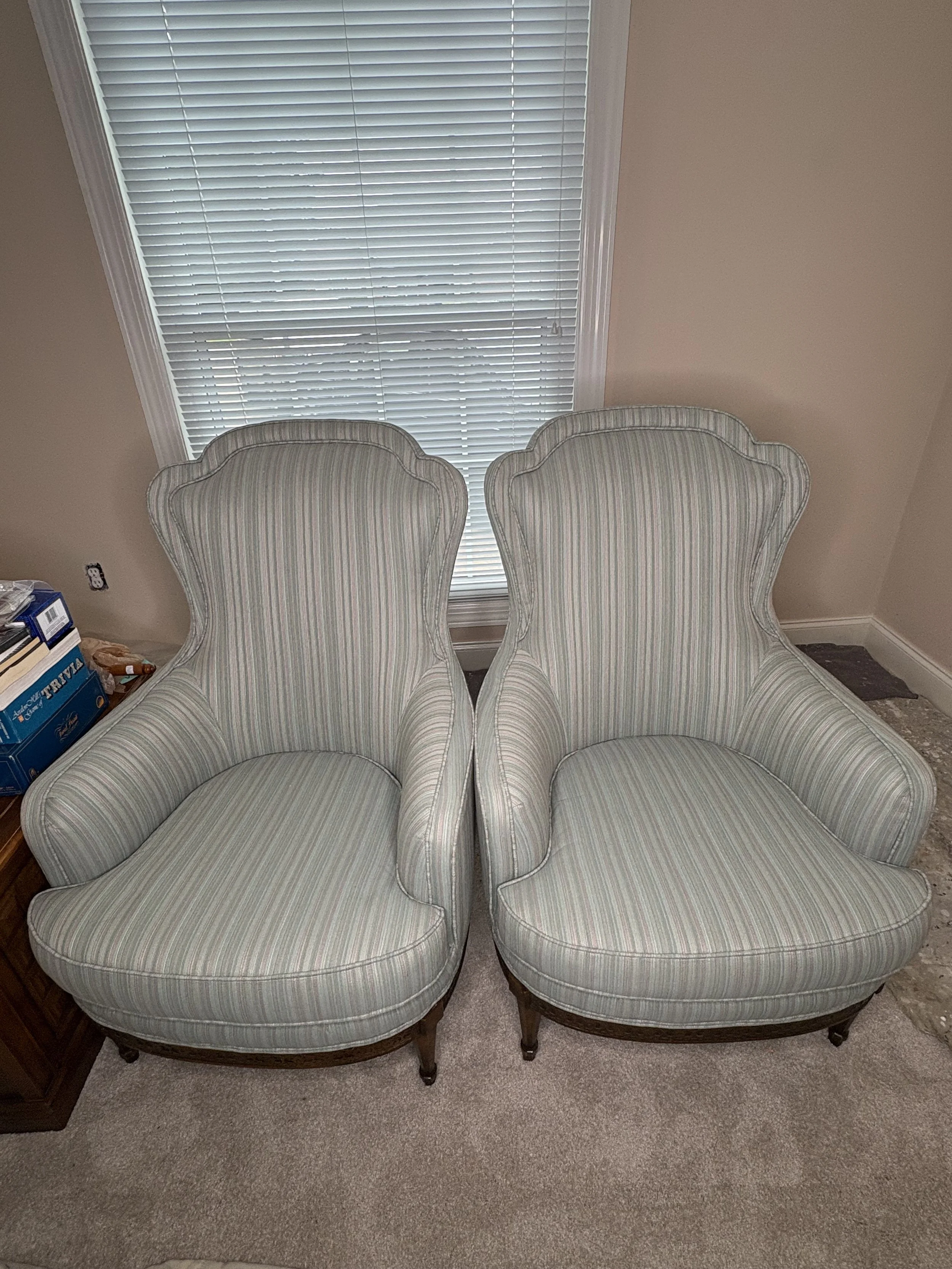 Two vintage armchairs with high, curved backs and striped upholstery, placed next to each other in a room with beige walls and a window with closed blinds.