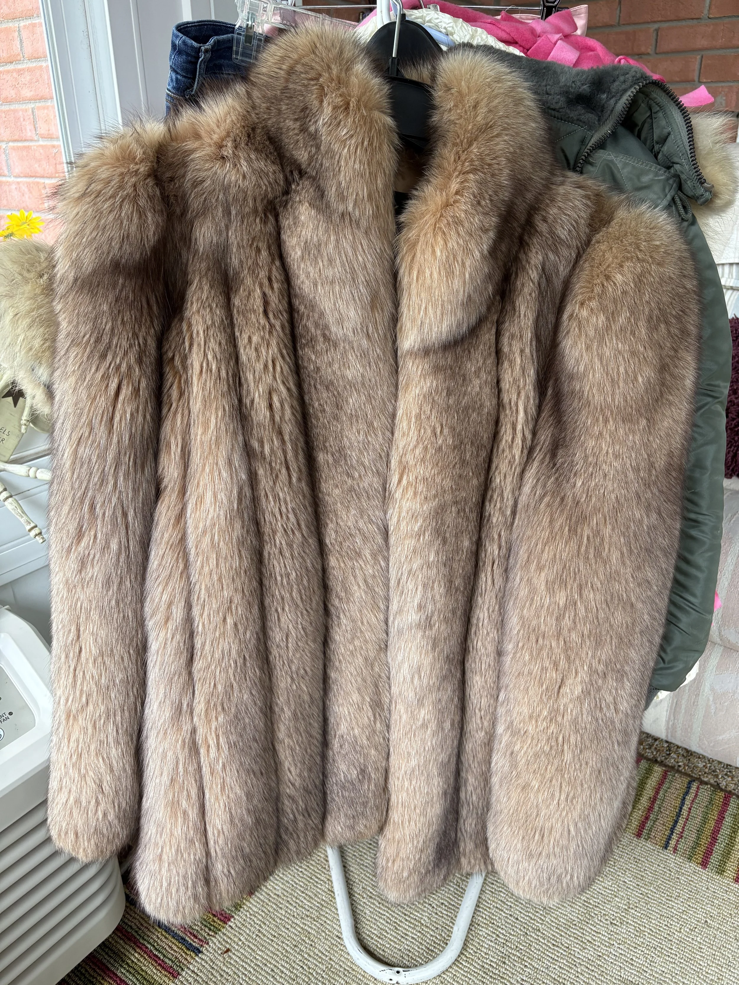 A brown fur coat hanging on a black hanger, displayed on a clothing rack, with jeans and a jacket in the background.