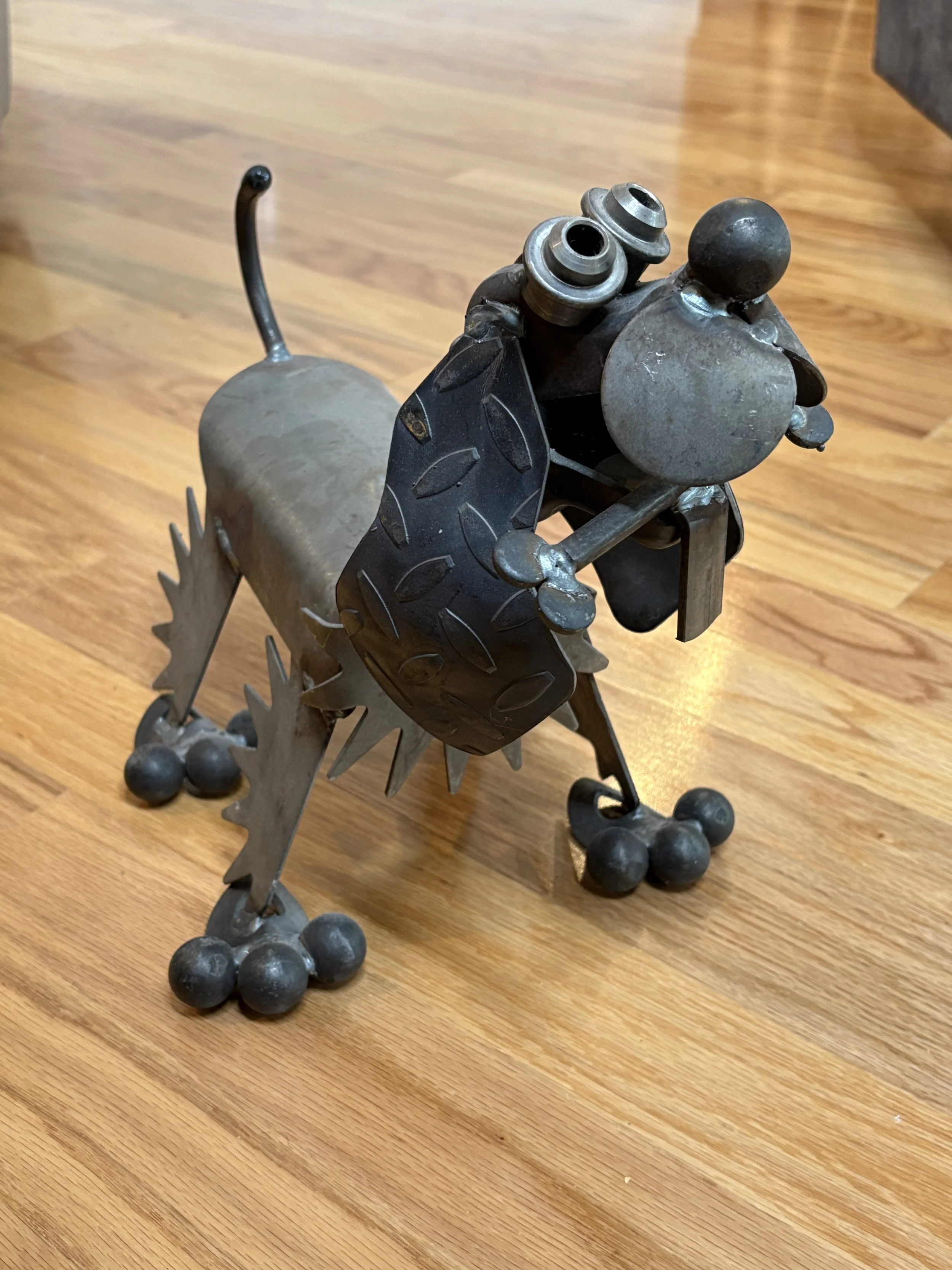 A sculpture of a dog made from metal parts, including gears, bolts, and plates, standing on a wooden floor.