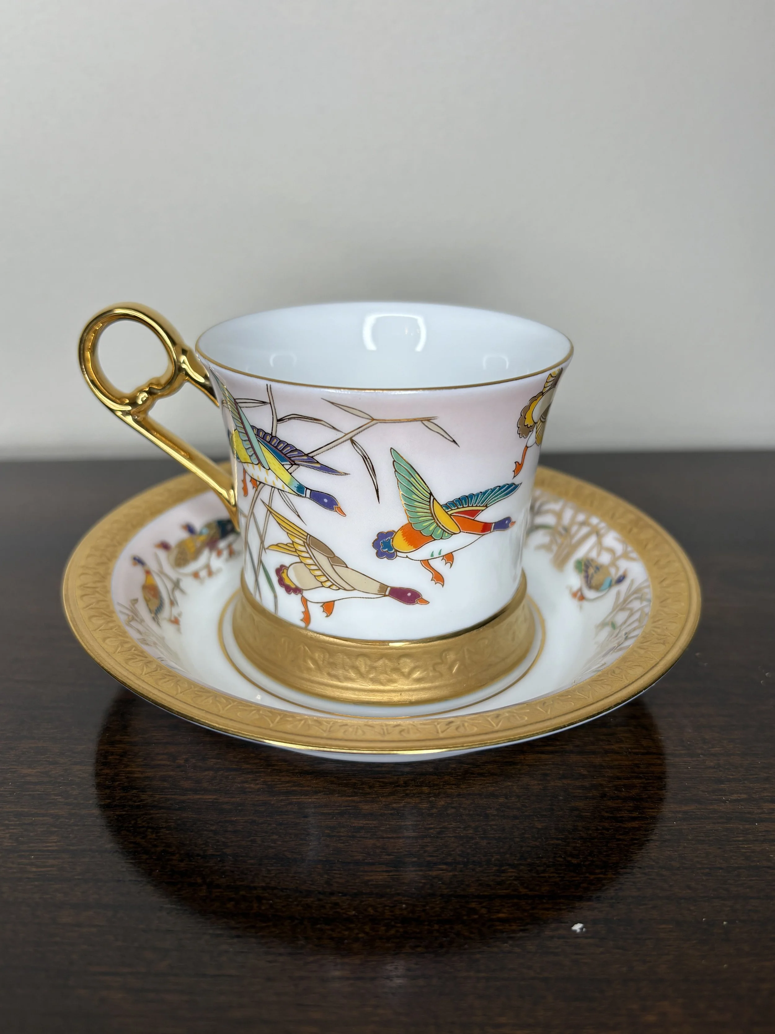 A decorative porcelain tea cup with a gold handle and a matching saucer, featuring colorful bird and nature designs, placed on a dark wooden surface.