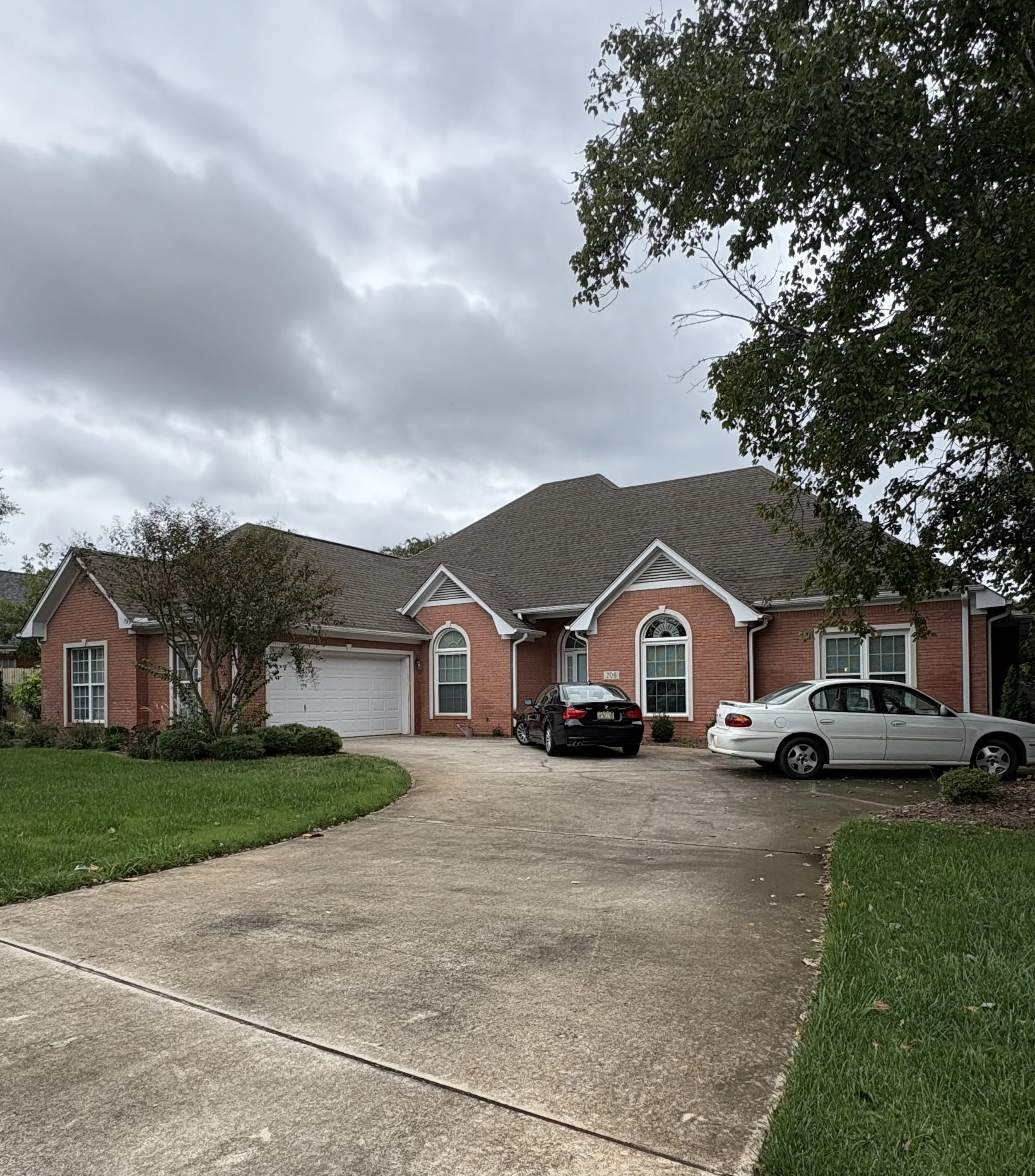 A brick house with a two-car garage, a driveway, and parked cars in the front yard under a cloudy sky, with trees and grass on the lawn.