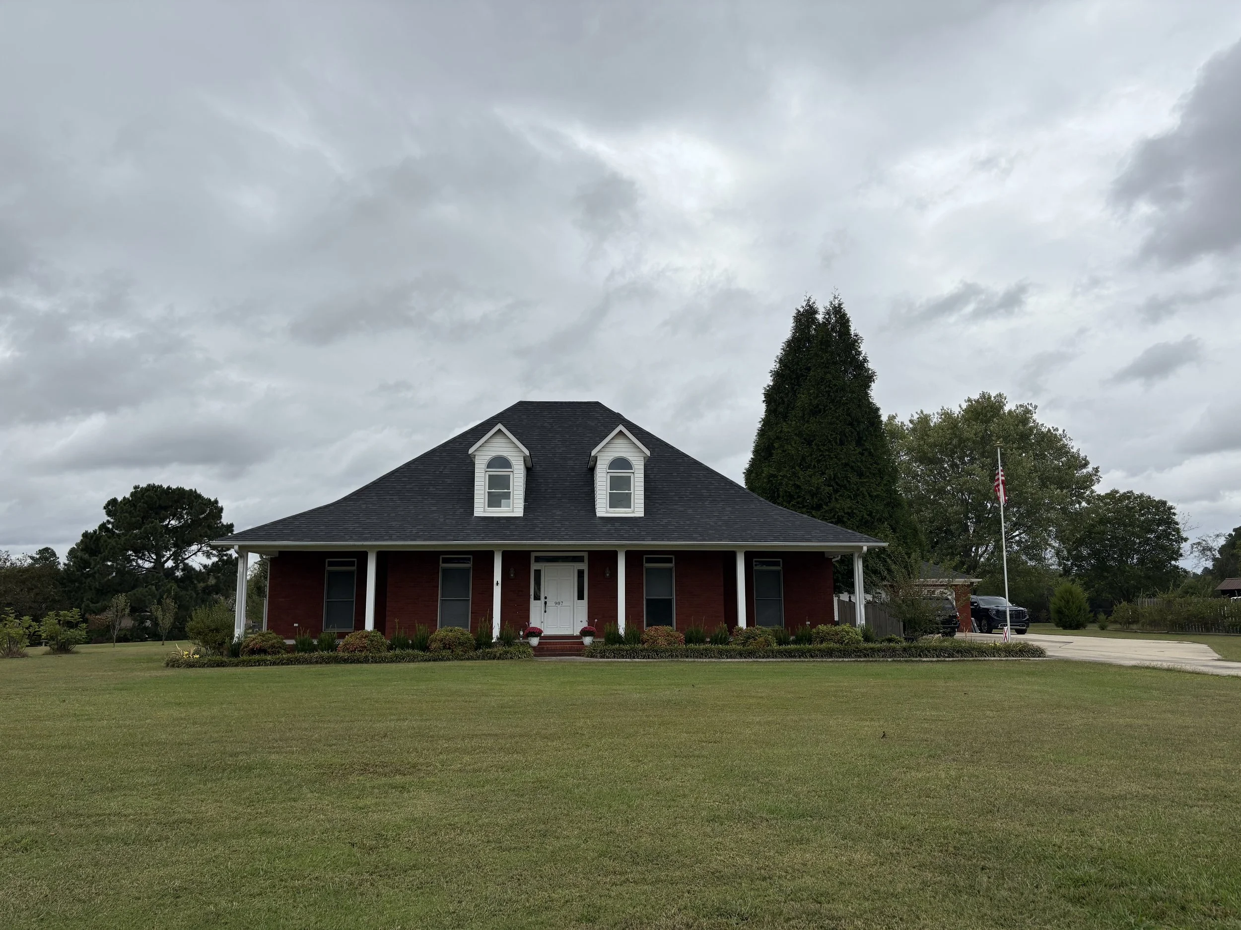 A large red brick house with a black shingle roof and two small dormer windows. The house has a white front door, a porch with four white columns, and landscaping with bushes and flowers in front. There is a grassy lawn, trees, and an American flag o