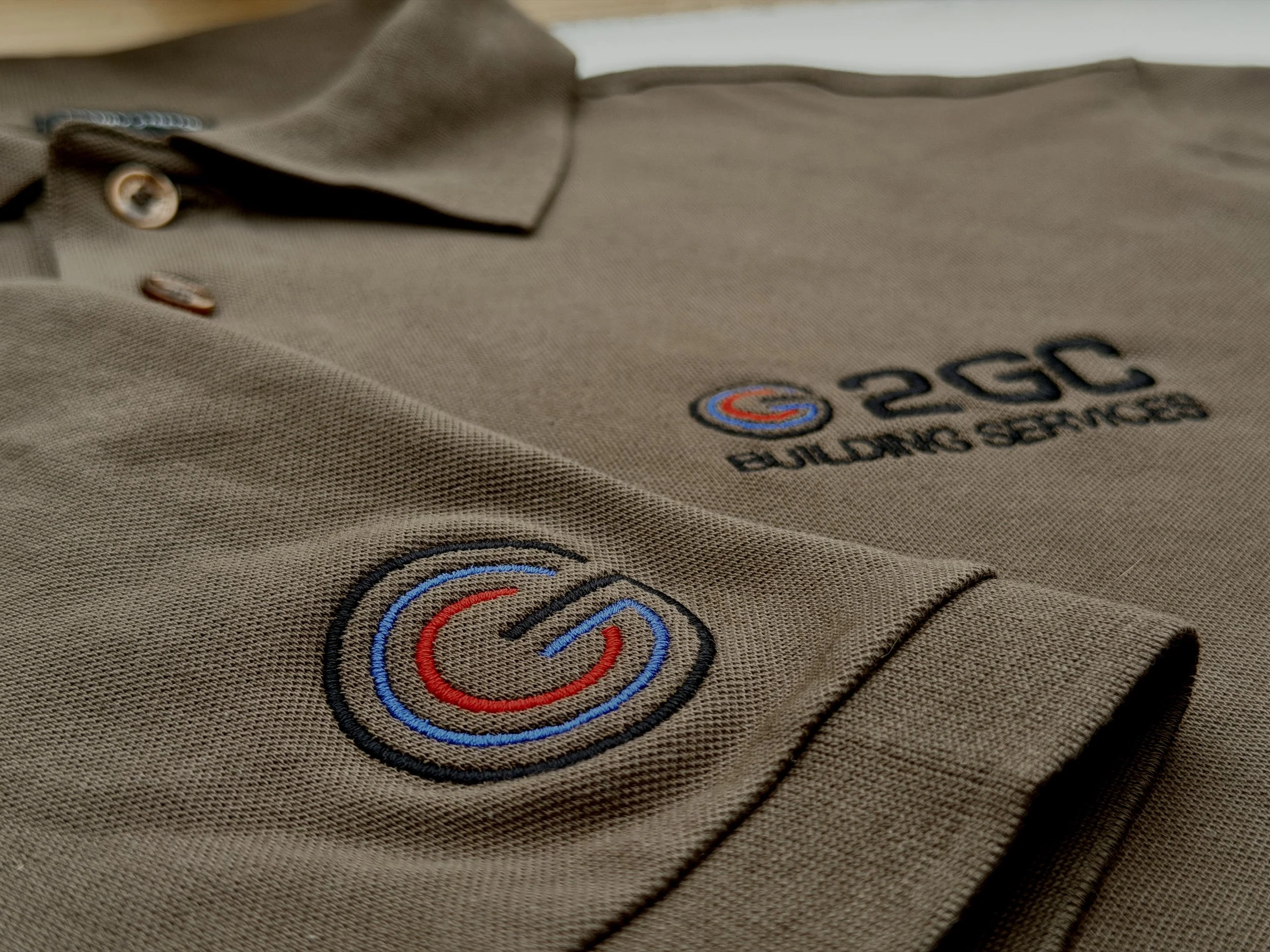 A brown polo shirt with an embroidered logo of a circular design in red, blue, and black, and text reading '2CC BUILDING SERVICES' on the chest.