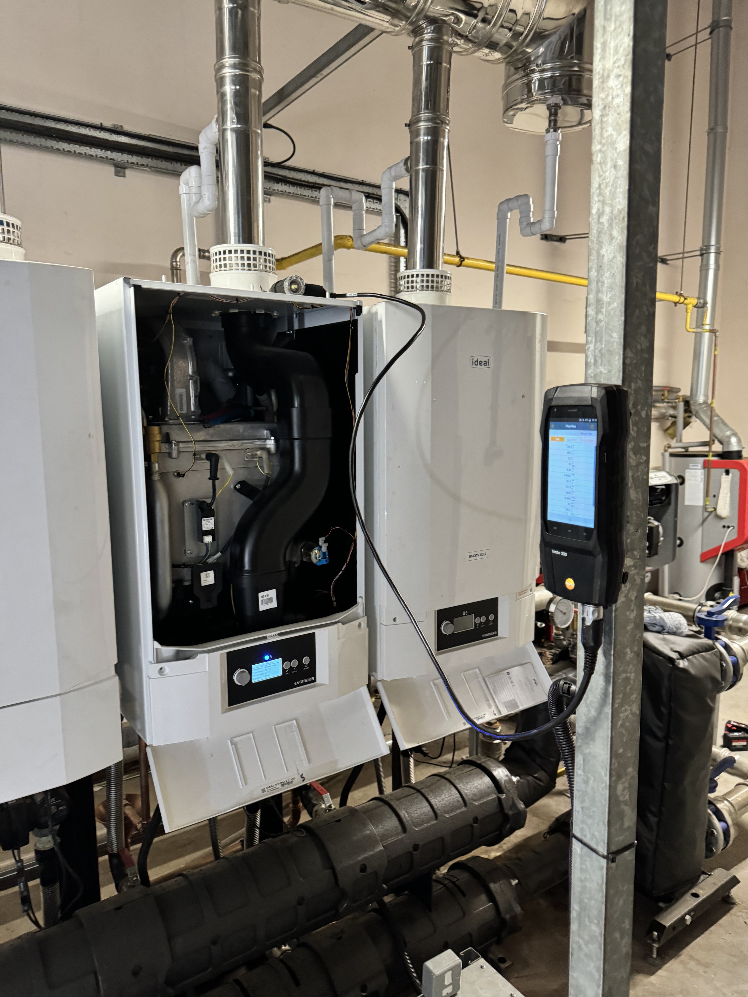 Industrial equipment with pipes, two white control units, and a handheld device attached by a cable, installed in a building utility room.