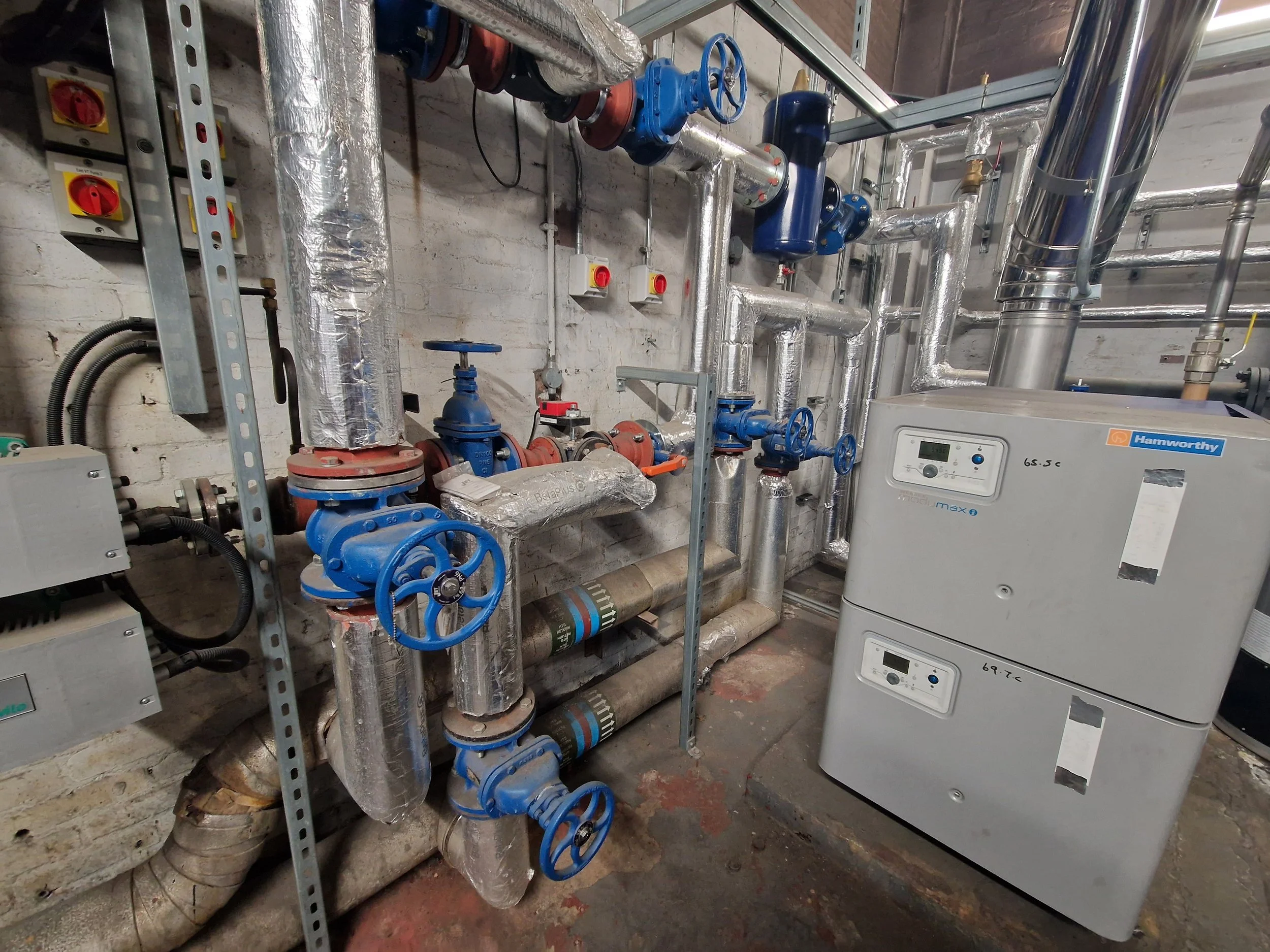 A mechanical room with pipes, blue valves, and control units, including a white boiler labeled 'Hamworthy' and labeled temperature settings.