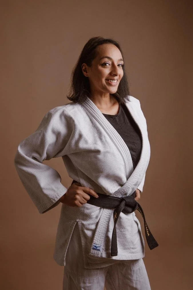 Femme souriante en kimono de judo blanc avec une ceinture noire, posant de profil contre un fond beige.