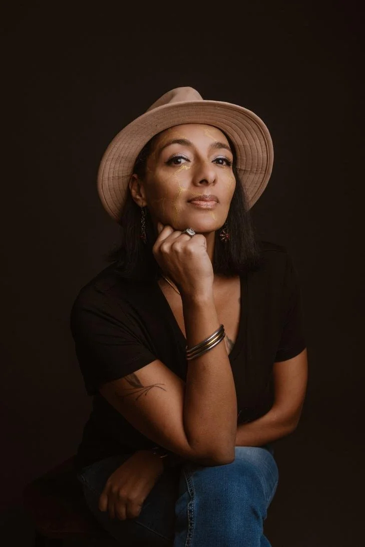 Portrait d'une femme portant un chapeau beige, vêtue d'un t-shirt noir, avec des bijoux, assise contre un fond sombre.