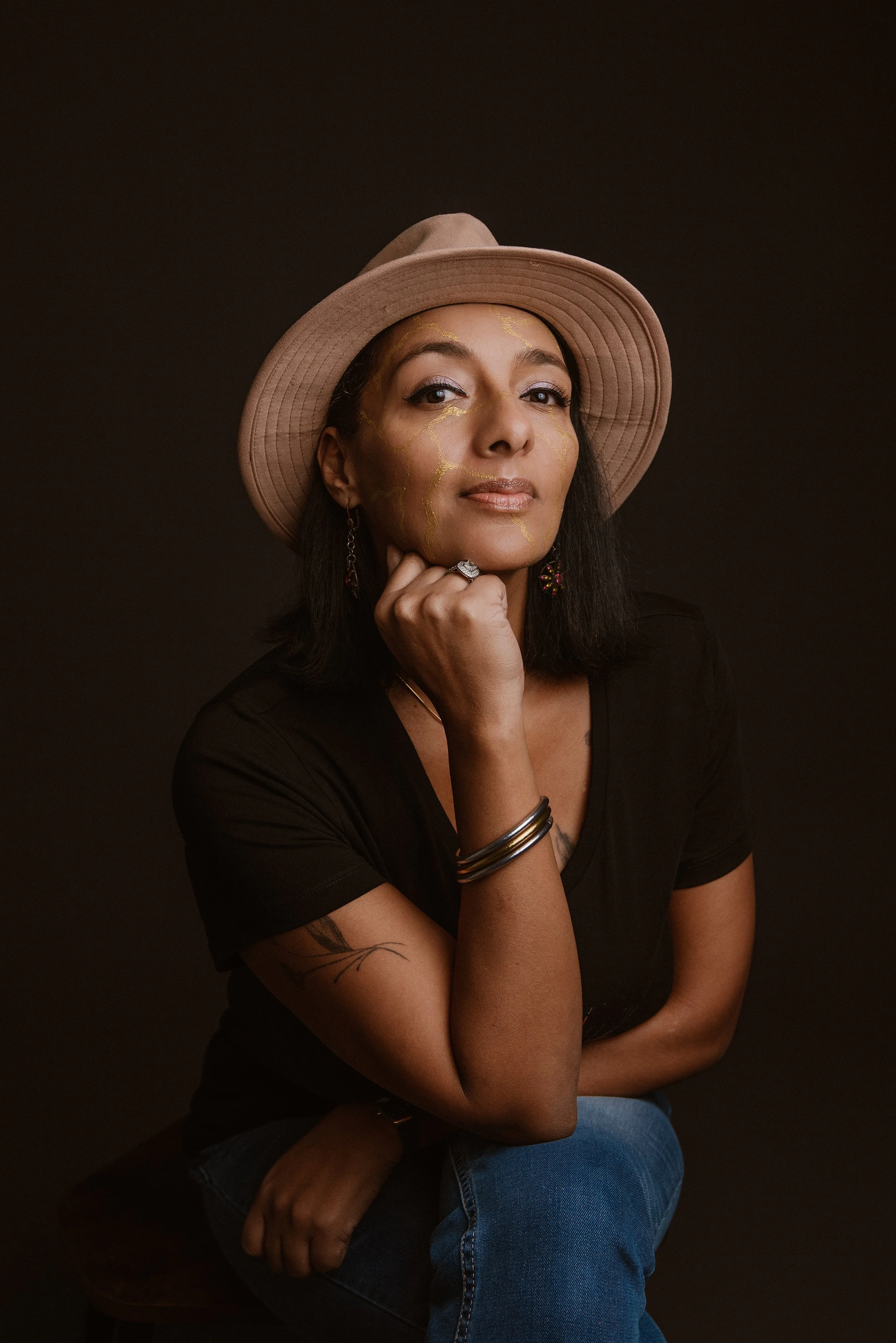 Angelique regarde droit dans les yeux, porte un chapeau beige, un tshirt noir et un jeans. Des traces de kintsugi sur son visage