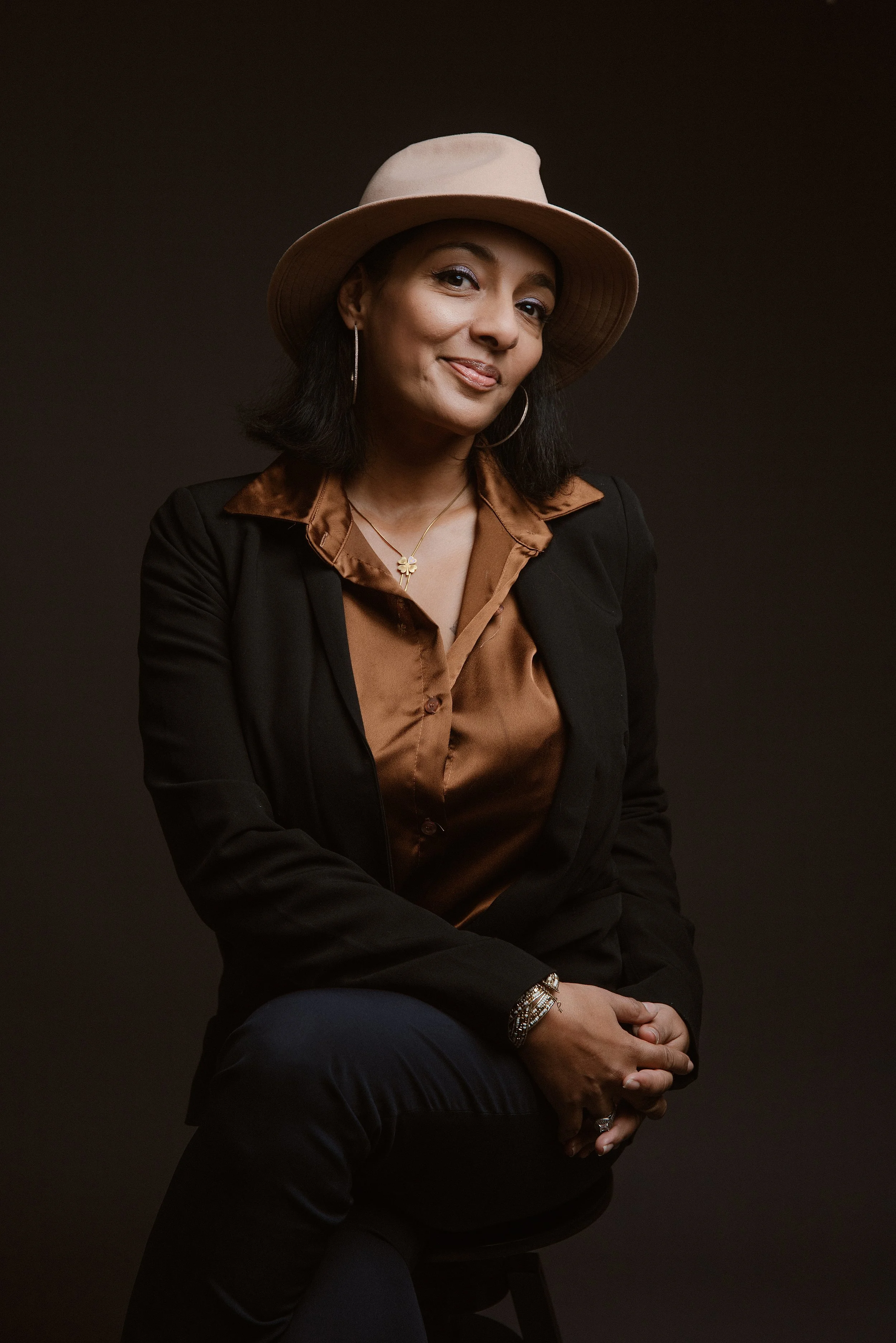 Une femme assise portant un chapeau beige, une chemise marron satinée, un blazer noir, des boucles d'oreilles et des bracelets, posant contre un fond sombre.