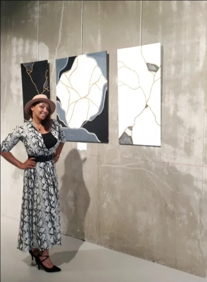 Angélique Tefenkgi dans une gallerie d'art, devant un tryptique de peintures représentant le Kintsugi. Elle porte une robe noire et blanche longue et un chapeau beige.