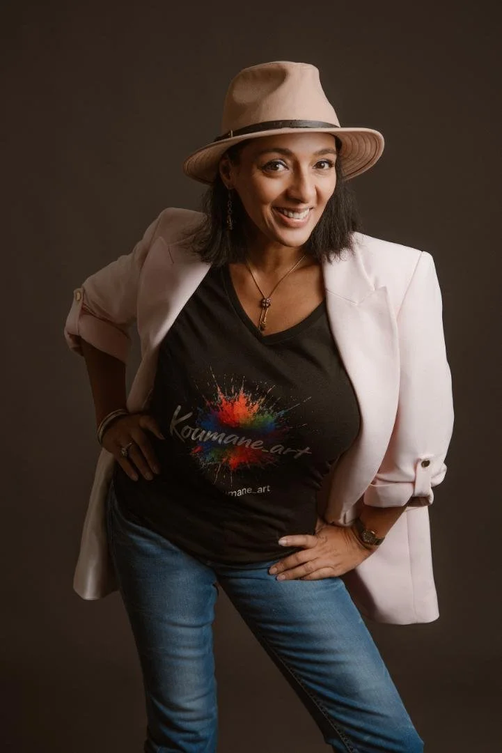 Femme souriante portant un chapeau beige, un blazer rose pâle, un t-shirt noir avec un logo coloré, et des jeans, debout contre un fond neutre.