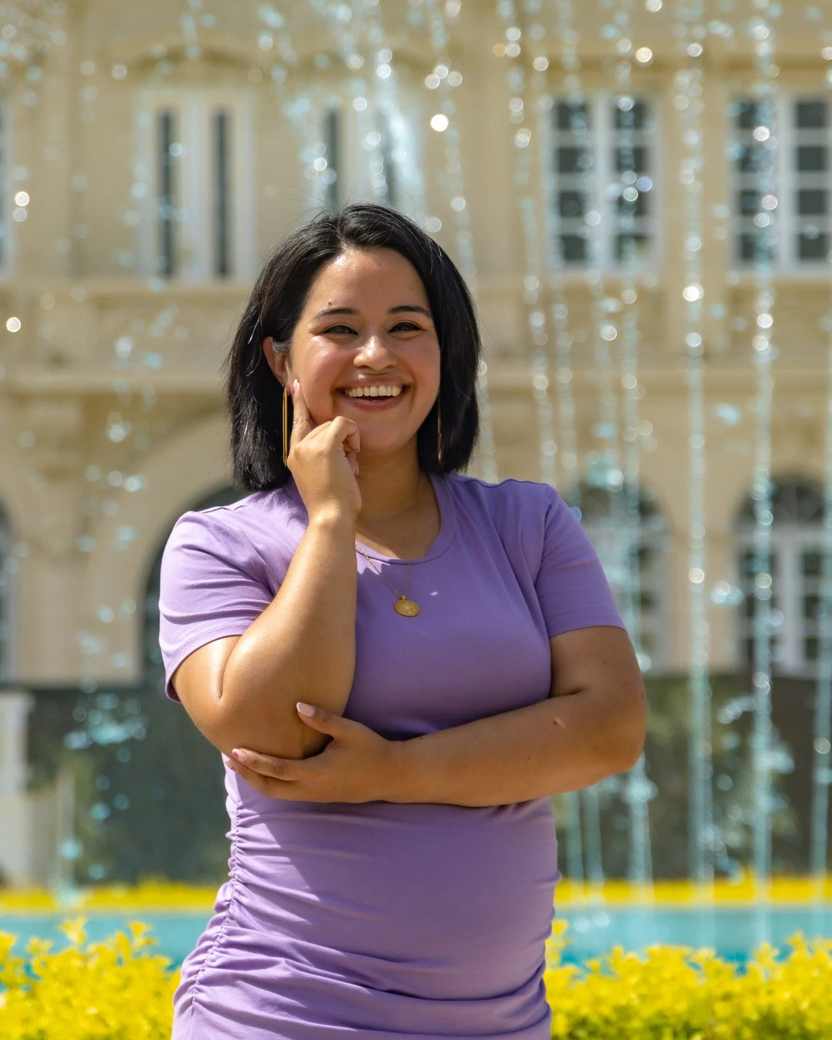 A woman with dark hair, smiling and posing with her hand on her face, wearing a purple top and a gold necklace, in front of a decorative fountain with water and glass beads.
