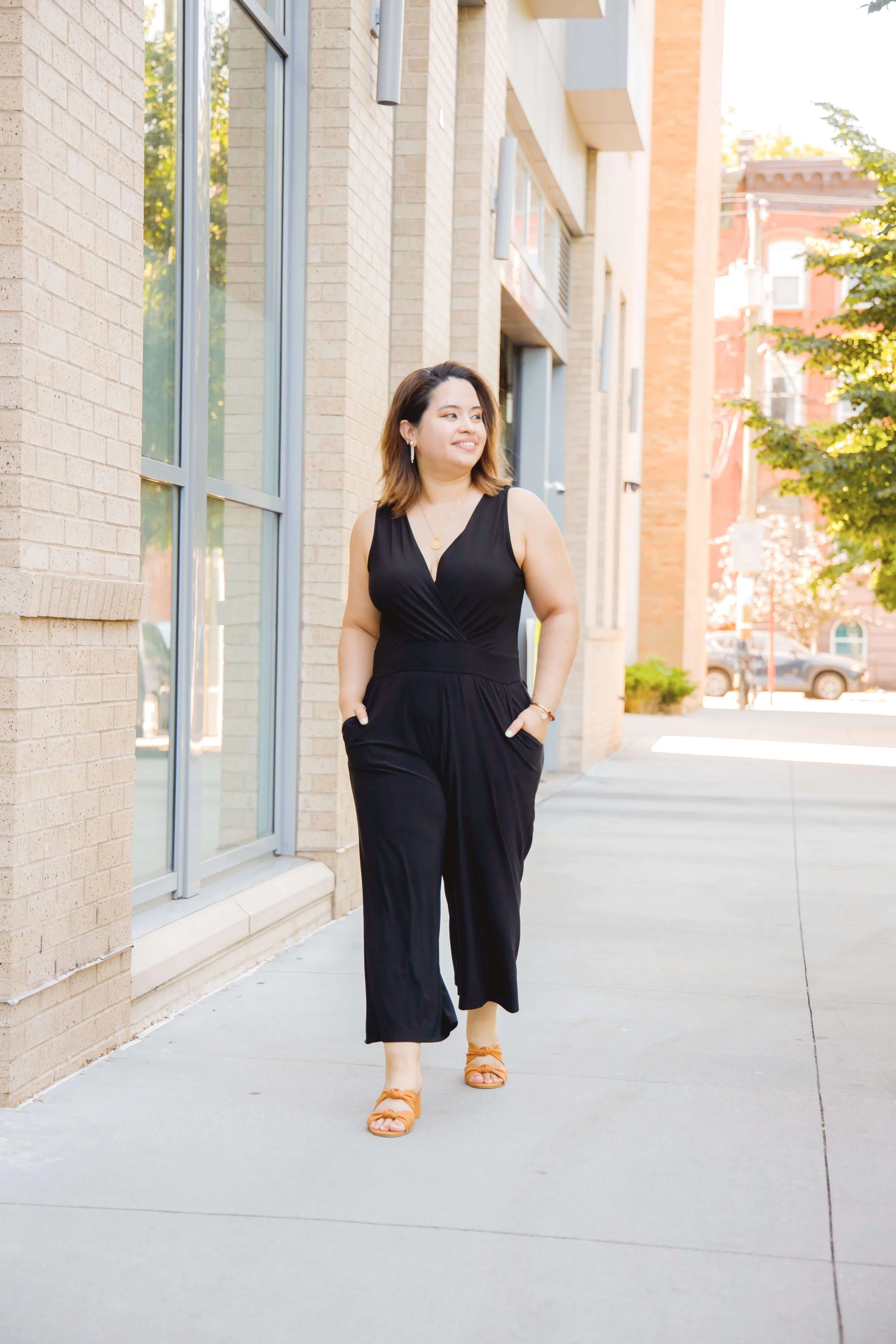 Geri Lopez, New Jersey trauma therapist for first generation women of color and queer adults, wearing a black jumpsuit and orange sandals walking on a city sidewalk next to brick buildings, with trees and parked cars in the background.