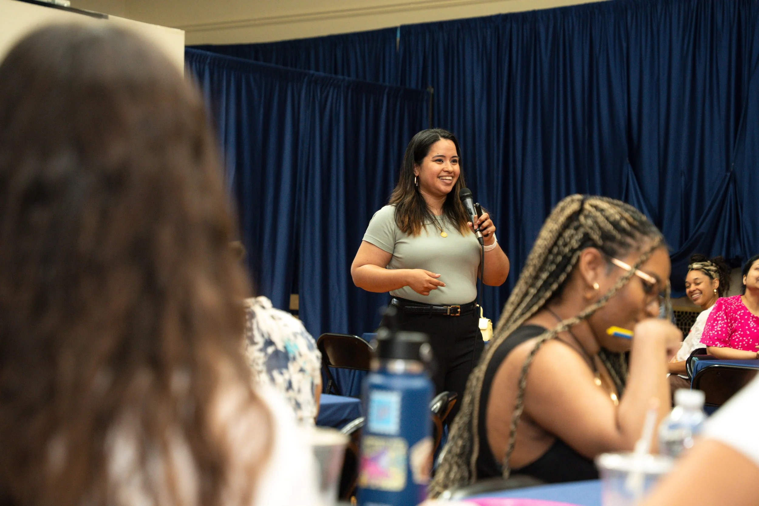 Geri Lopez, New Jersey trauma therapist standing and smiling while holding a microphone in front of a dark blue curtain, with several women seated at tables in the background during an indoor event.