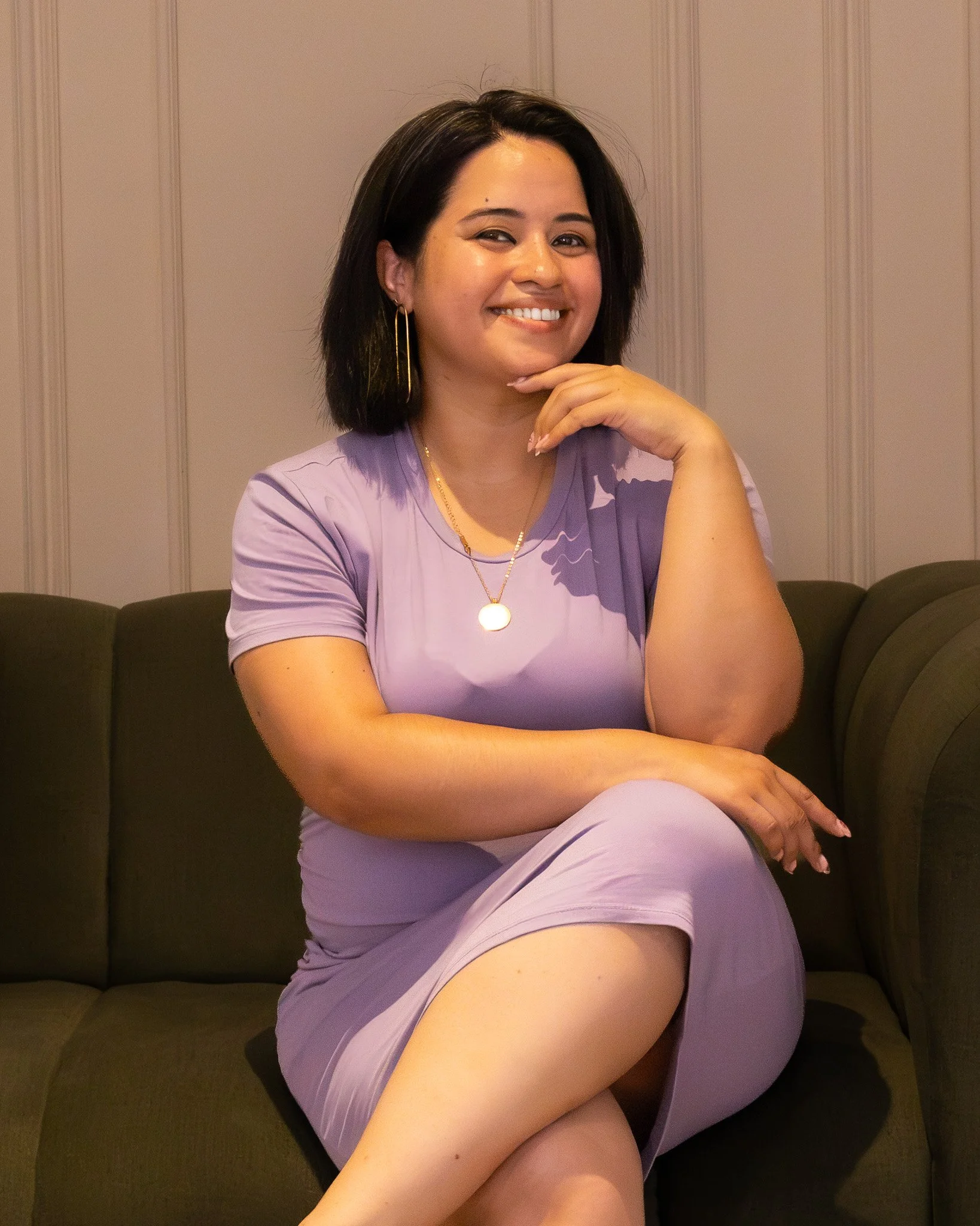 A smiling woman with dark hair, wearing a lavender top and jewelry, sitting on a brown couch with paneled walls in the background.