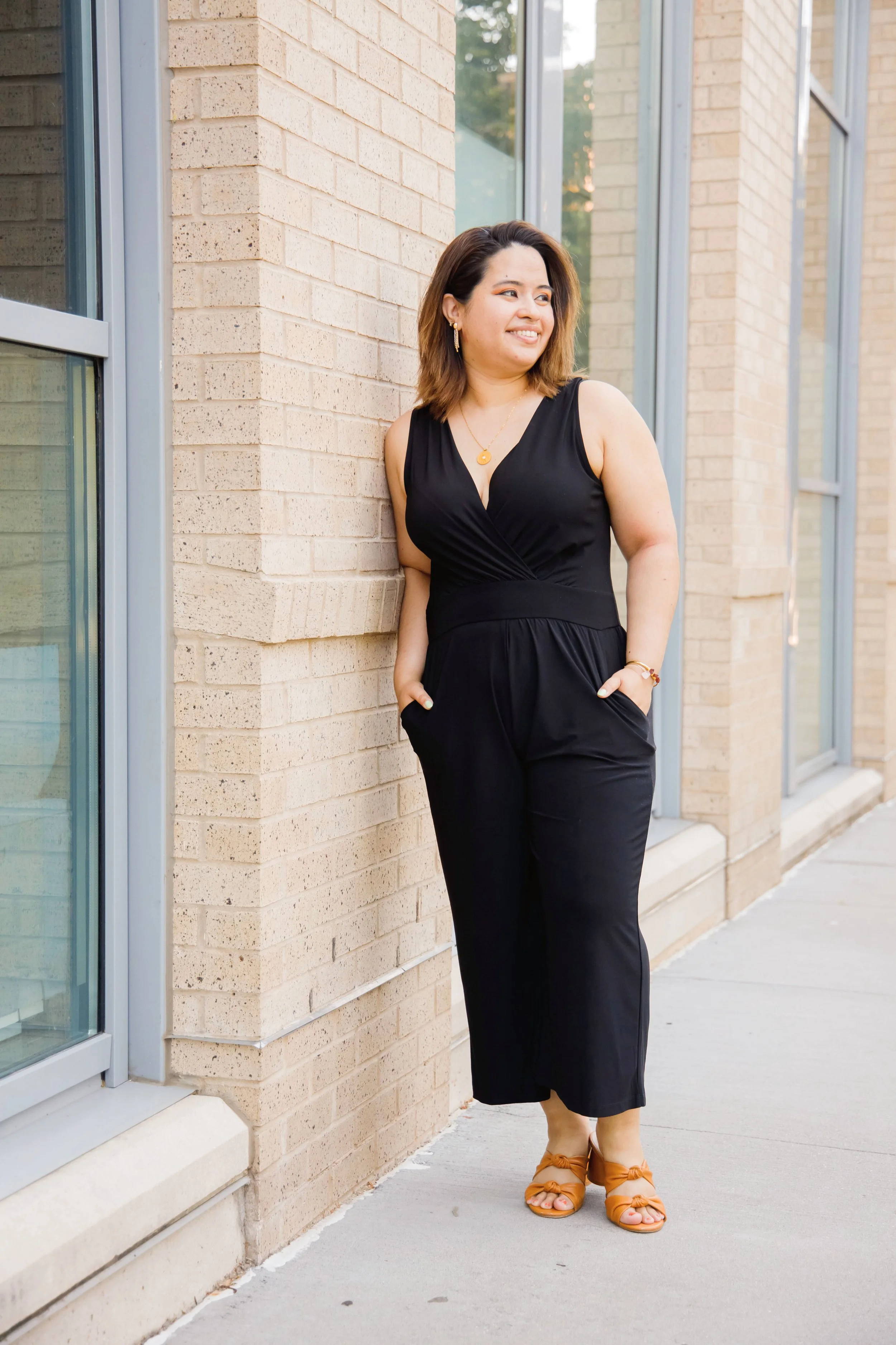 Geri Lopez, New Jersey trauma therapist with shoulder-length brown hair wearing a black sleeveless jumpsuit and tan sandals, standing next to a brick wall with large windows, smiling and looking to her left.
