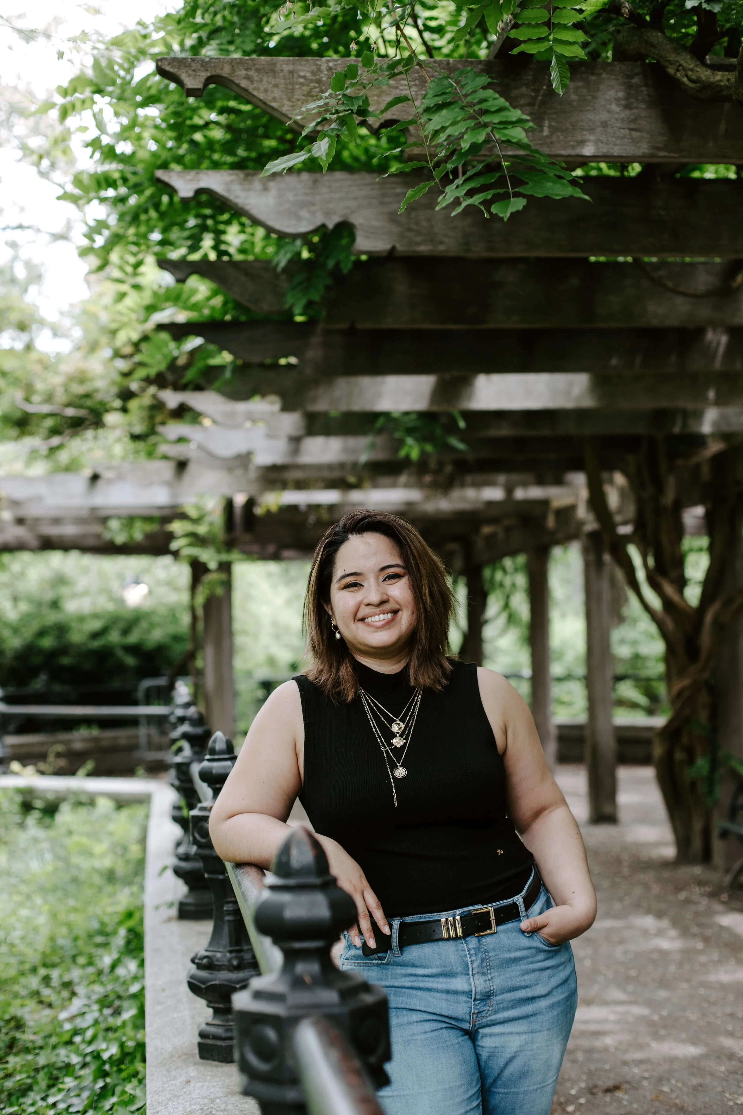 Geri Lopez, New Jersey trauma therapist for first generation women of color and queer adults,  shoulder-length brown hair, wearing a black sleeveless top and layered necklaces, smiling while sitting on a park bench under a pergola with green foliage.