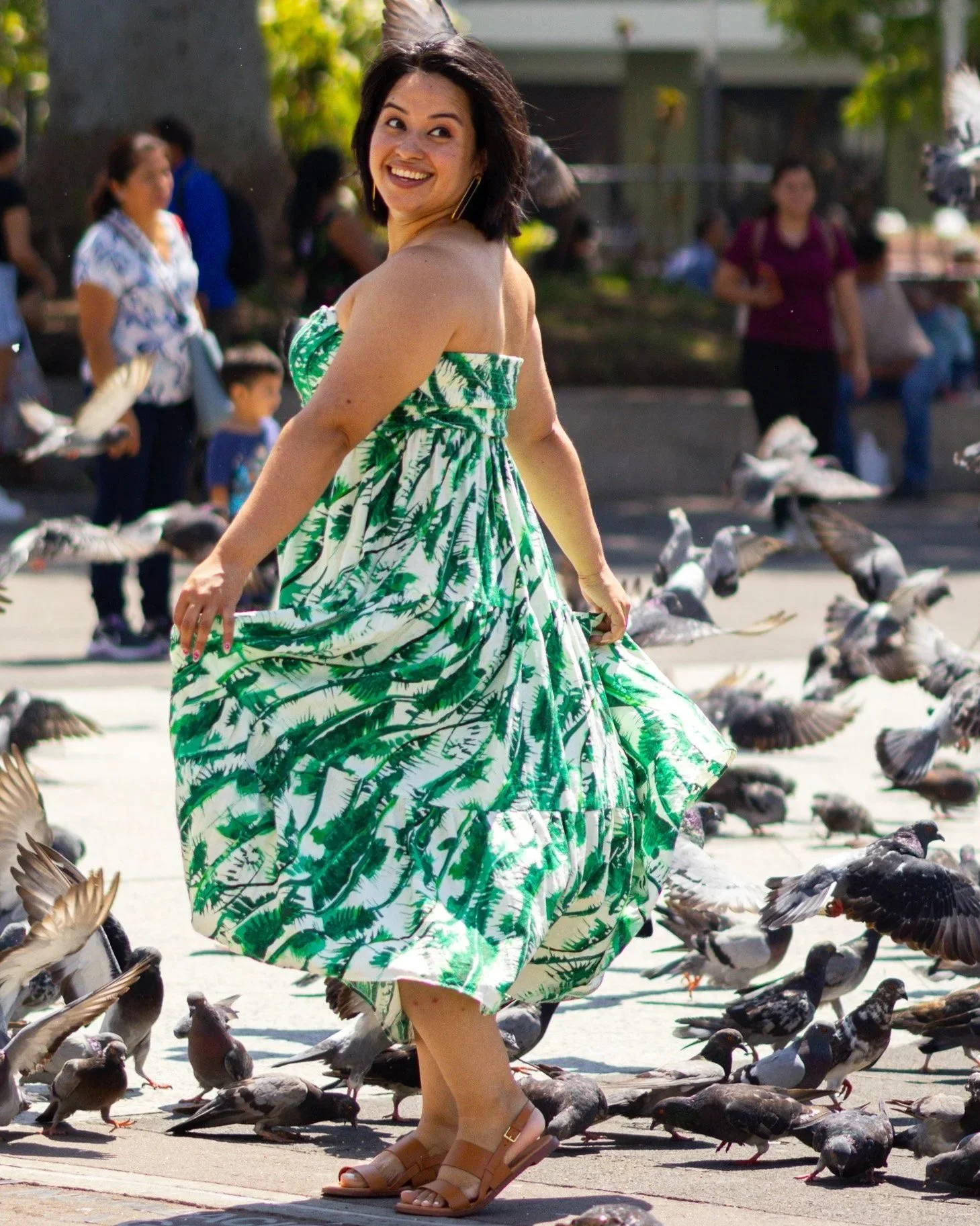 A woman in a green and white leafy dress smiling and spinning among pigeons in a park.