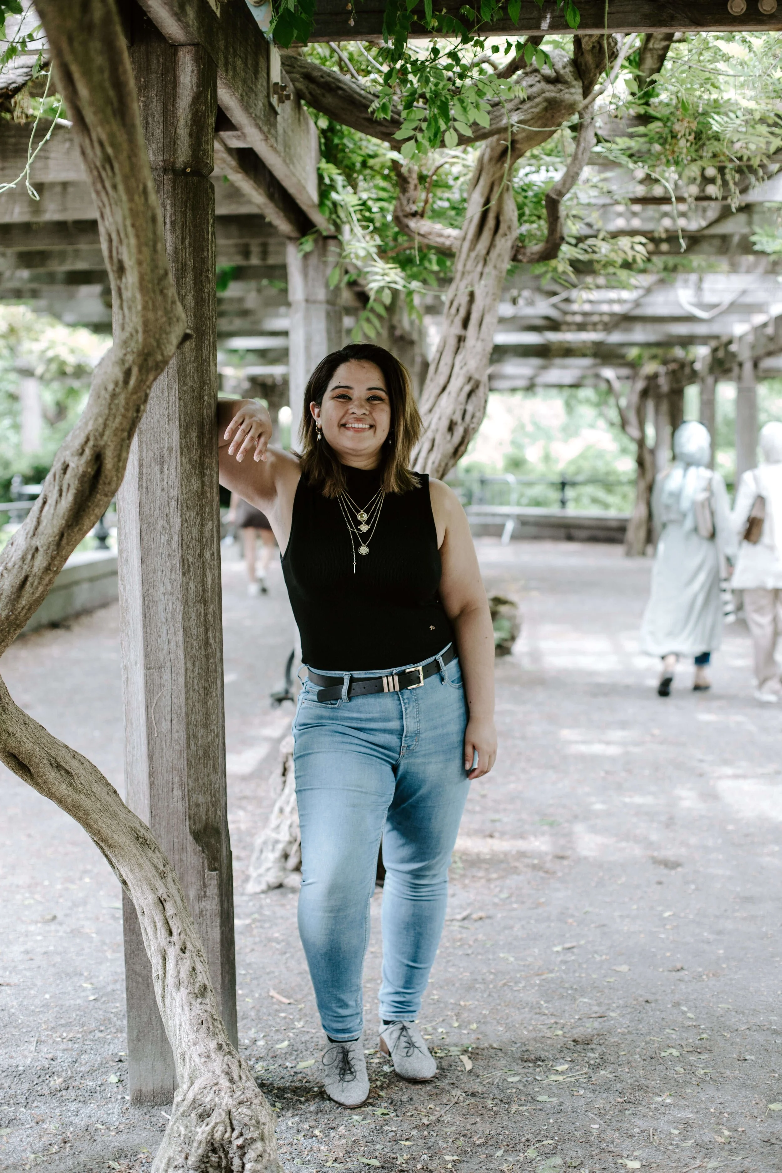 Geri Lopez, New Jersey trauma therapist with a shoulder-length brown hair, wearing a black sleeveless top, light blue jeans, and gray sneakers, is smiling and leaning against a wooden post under a pergola with trees in the background.