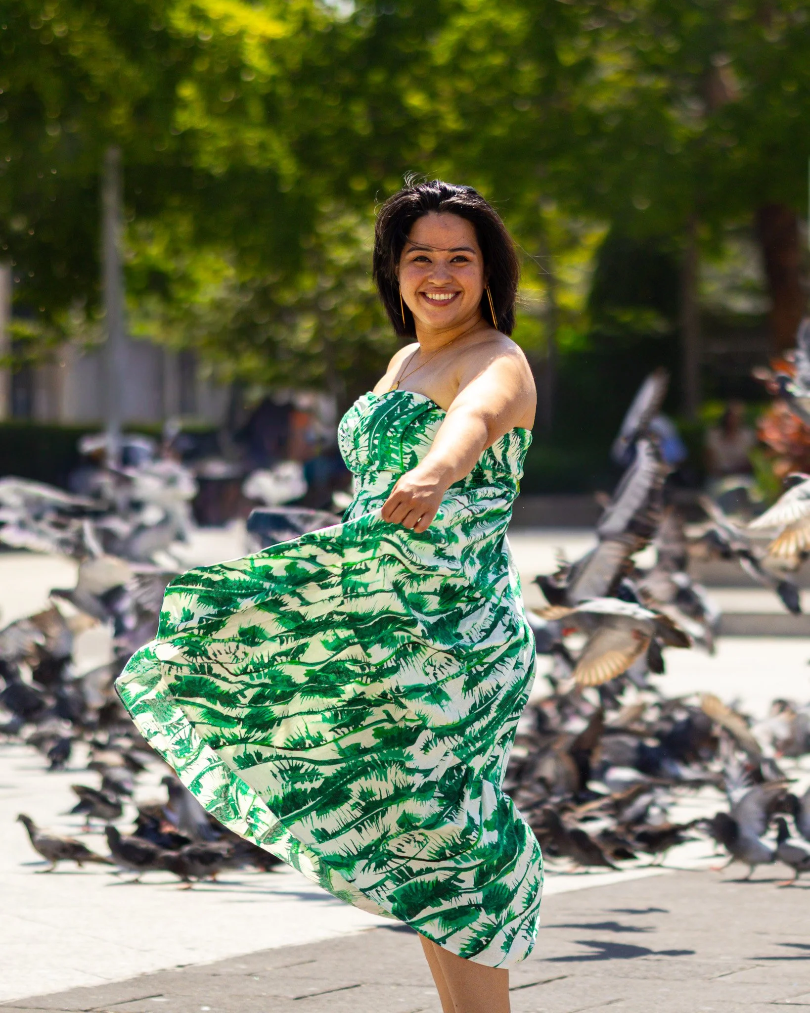 A smiling woman with black hair and light skin, wearing a green and white dress with tropical leaf patterns, is outdoors on a sunny day near a row of parked scooters and trees in the background.