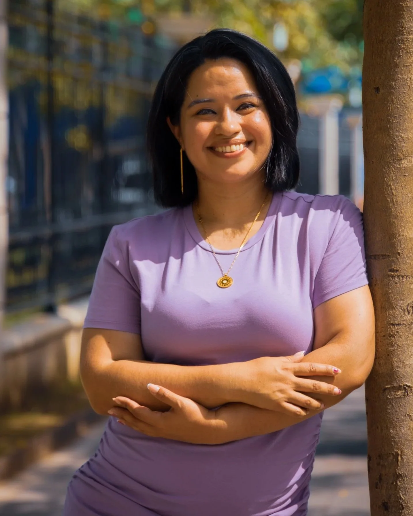 A woman with black hair, wearing a purple shirt and gold jewelry, standing outdoors next to a tree, smiling with arms crossed.
