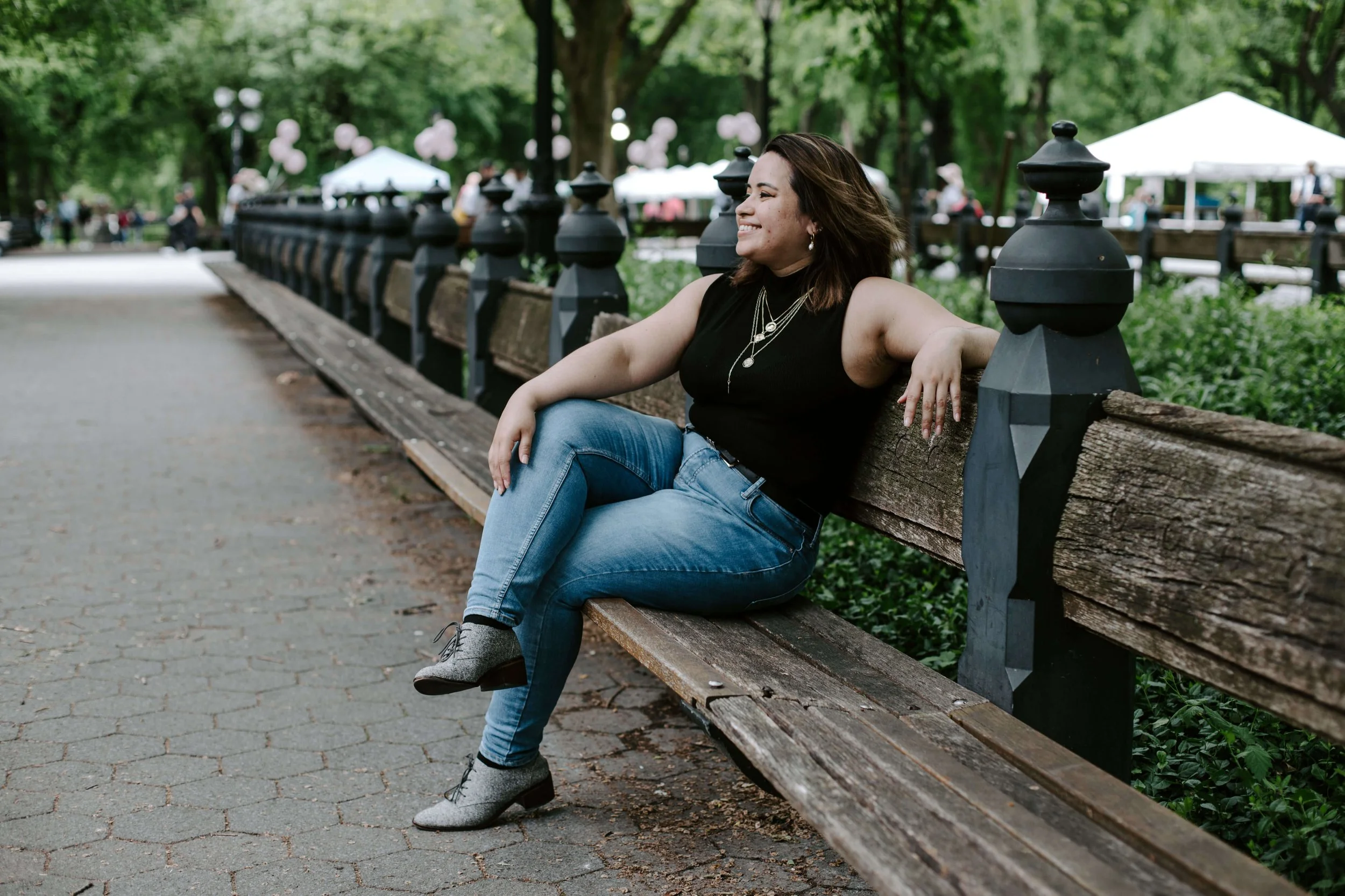 Geri Lopez, New Jersey trauma therapist for first generation women of color and queer adults, sitting on a park bench, smiling, with lush green trees in the background and white tents in the distance.