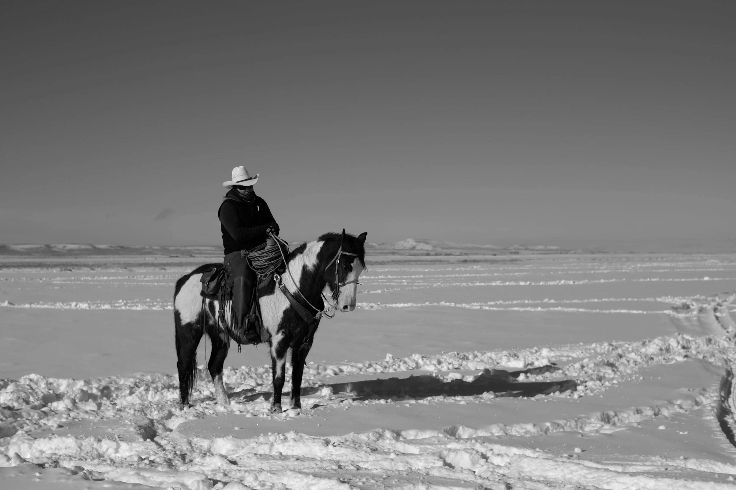 A person wearing a cowboy hat and dark clothing riding a pinto horse on snowy terrain with a vast, snow-covered landscape in the background.