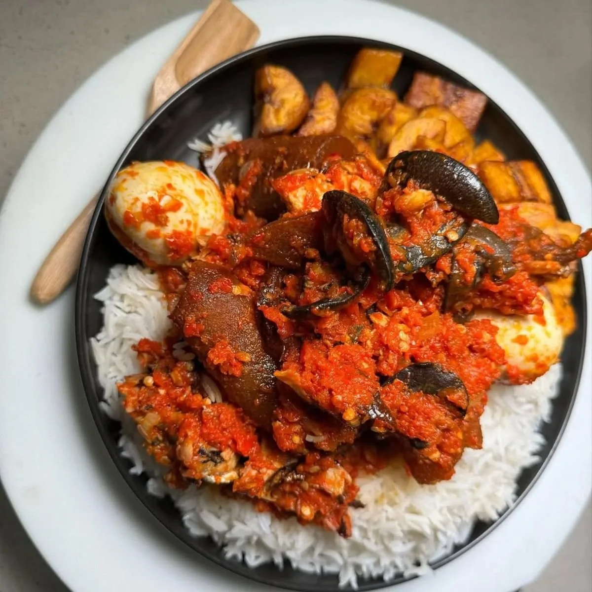 Plate of white rice topped with a snail pepper stew containing fish, cow skin, boiled egg, plantains, and fried fish, with a spoon on a white background.