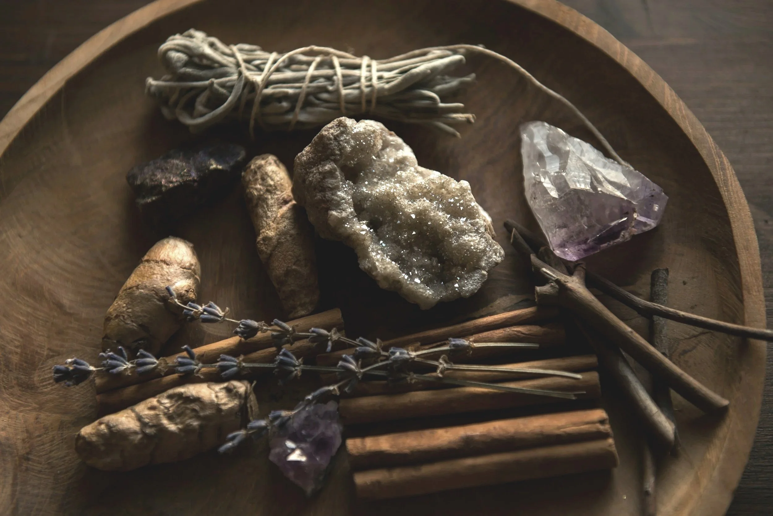 A wooden tray containing herbal and mineral objects, including lavender sprigs, cinnamon sticks, ginger roots, a raw amethyst gemstone, a large mineral specimen, and dried plant stems.