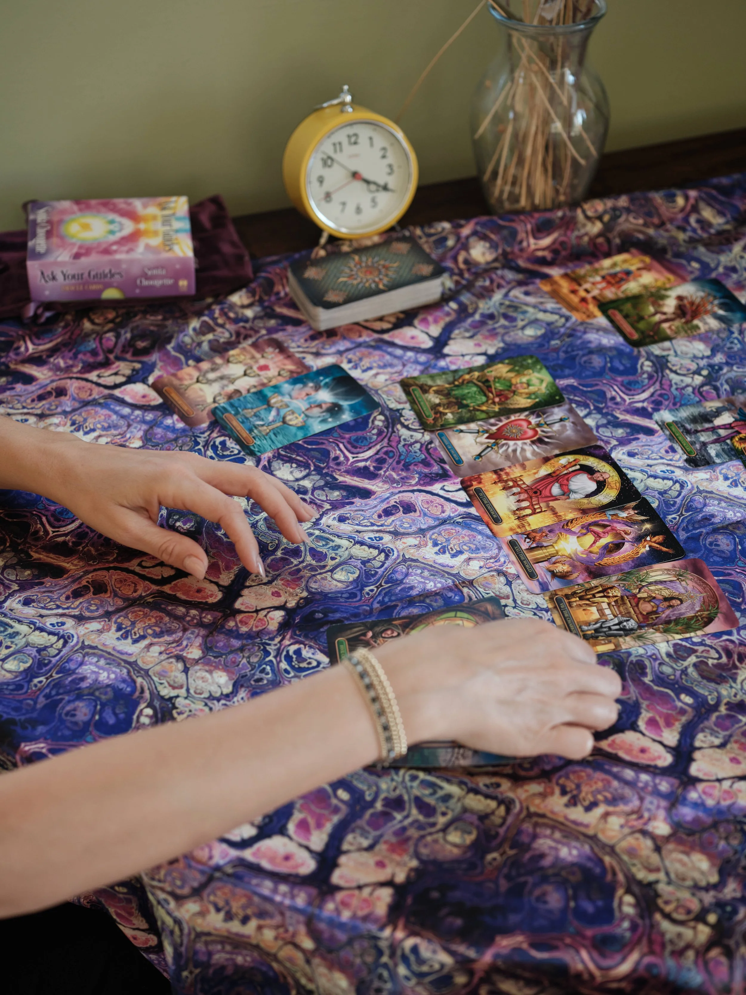 A person with a bracelet on their wrist reaches to the tarot cards laid out on a colorful, patterned cloth on a table. There are decks of cards and a tarot guidebook nearby, with a yellow clock and a glass vase with sticks in the background.
