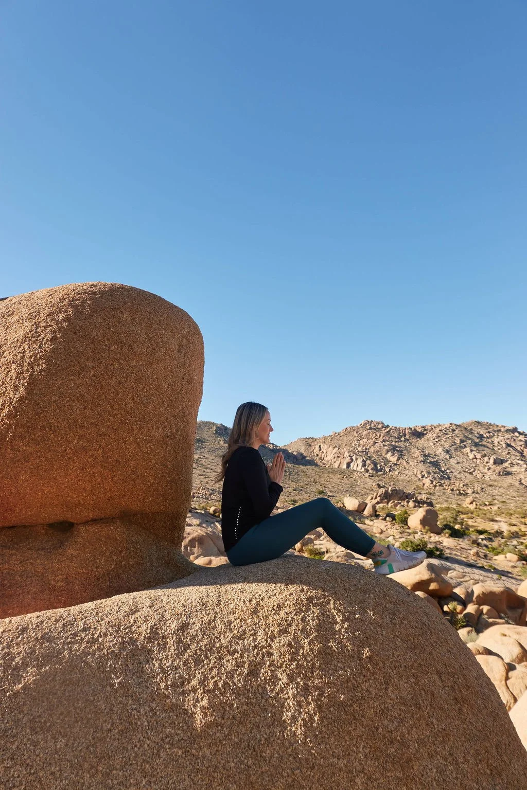 A woman practicing yoga in a desert landscape with large rocks and mountains in the background, under a clear blue sky.
