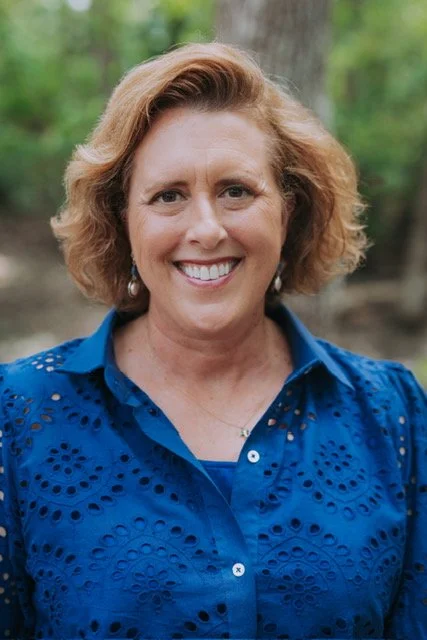 A smiling woman with light skin and short, wavy blonde hair, wearing a blue eyelet blouse, standing outdoors in a wooded area.