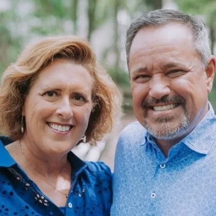 A smiling middle-aged woman with reddish-brown hair and a middle-aged man with gray hair and beard, both outdoors with trees in the background.