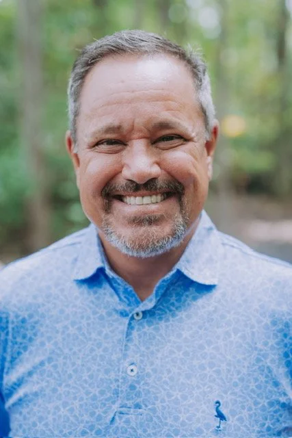 Smiling middle-aged man with short gray hair and a beard, wearing a blue patterned button-up shirt, outdoors in a green, wooded area.