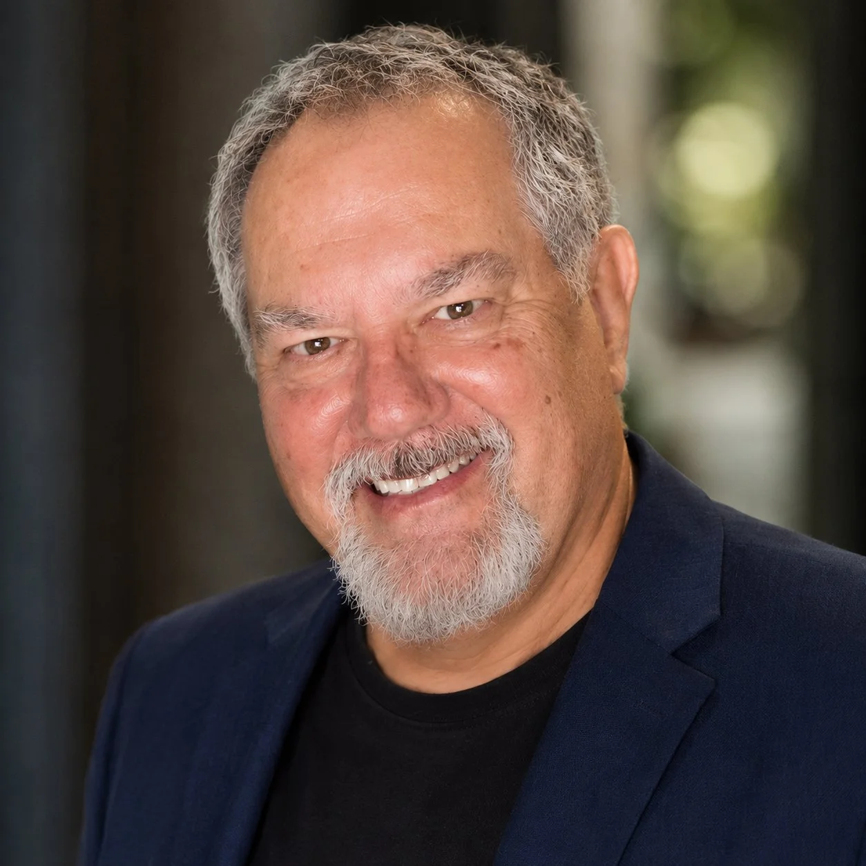 Headshot of a smiling middle-aged man with Salt and Pepper hair, a beard, and mustache, wearing a dark blue blazer and black shirt, against a blurred background.
