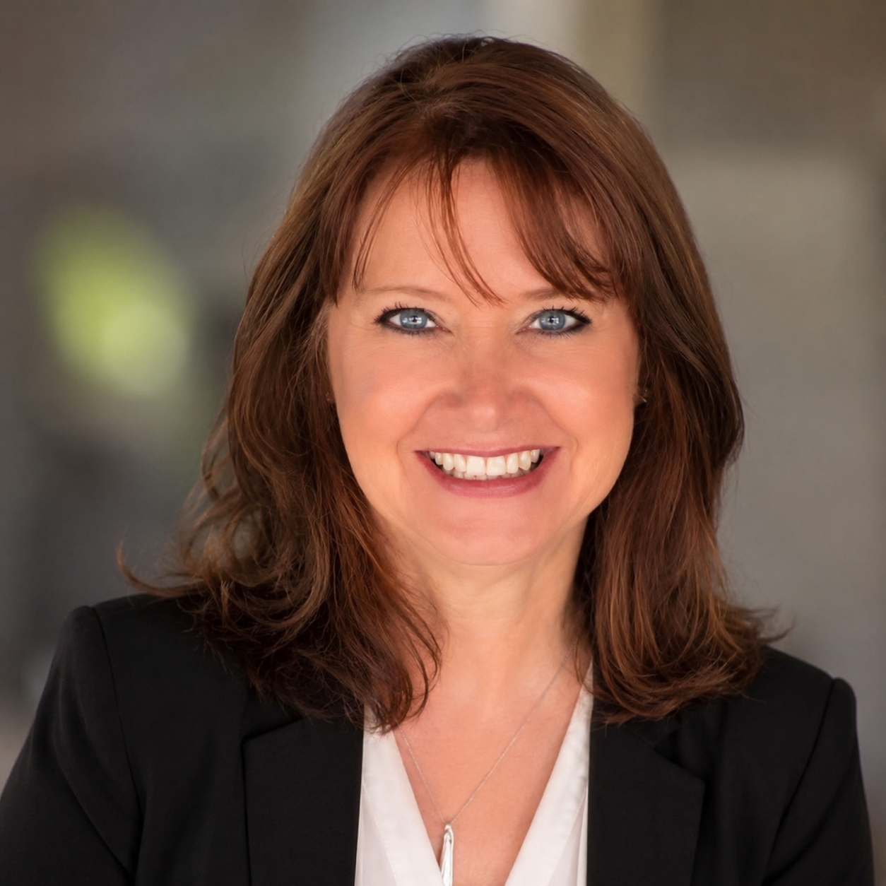 A woman with shoulder-length auburn hair smiling, wearing a black blazer and a white blouse, against a blurred indoor background.
