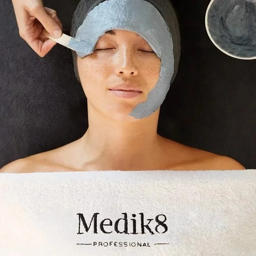 A woman with freckles relaxes with her eyes closed at a spa or skincare salon, receiving a facial treatment with a gray facial mask or peel applied to her face, and a person gently removing part of the mask with a spatula. There are small bowls with skincare products nearby.