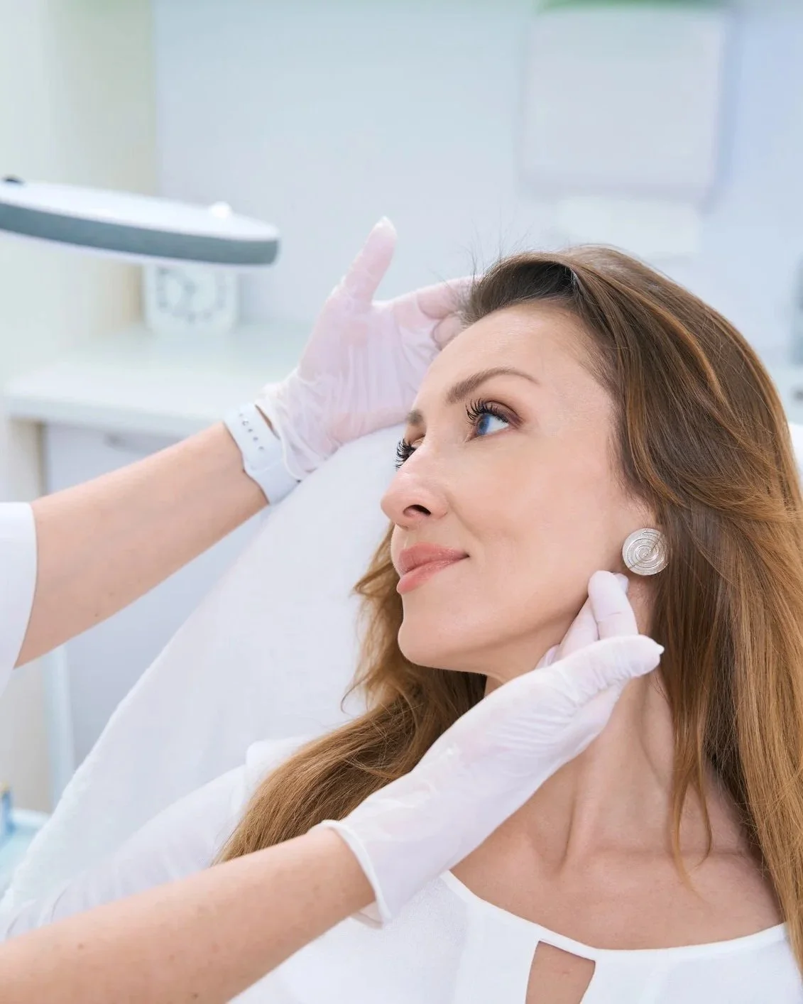 A woman with long red hair receives a cosmetic or dermatological treatment from a healthcare professional in a clinical setting.